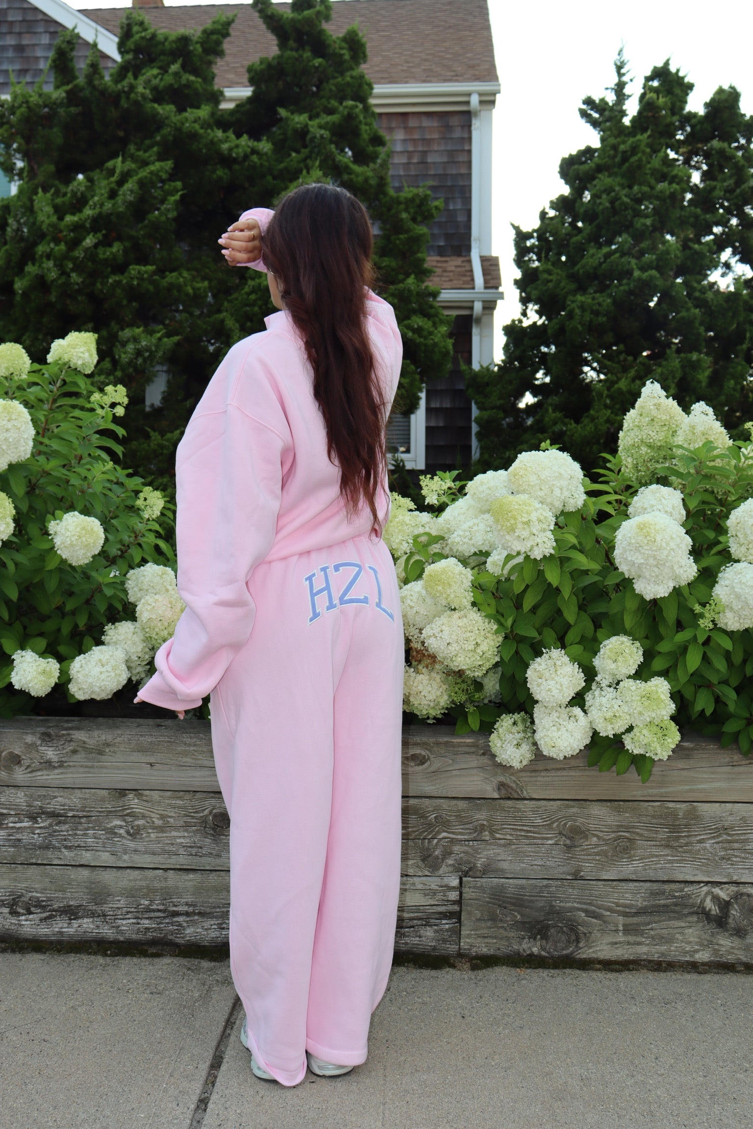 Person wearing pink HZL sweatpants beside blooming white hydrangeas outside a home