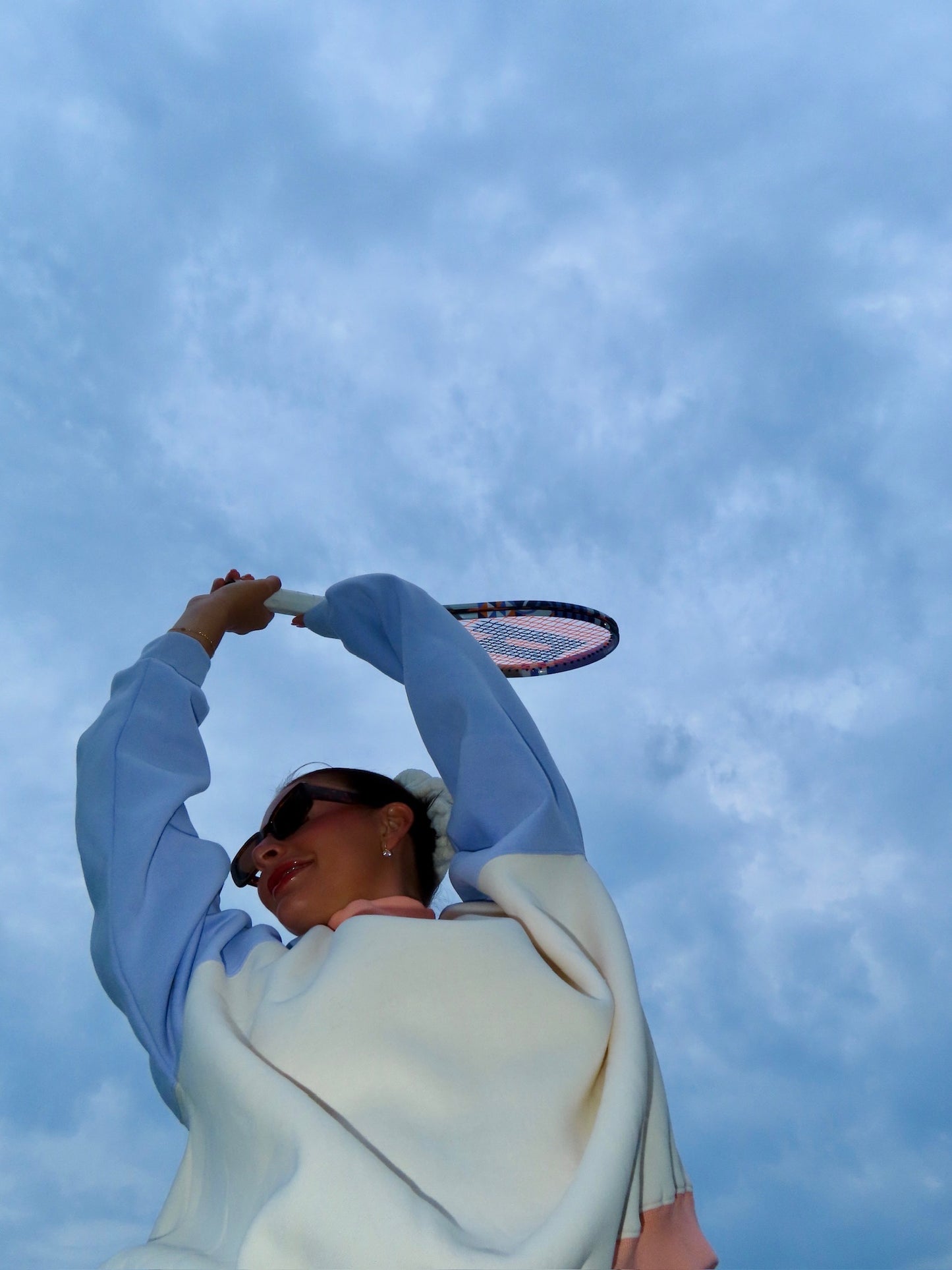 Person wearing a light-colored mockneck sweatshirt holding a tennis racket against a cloudy sky background