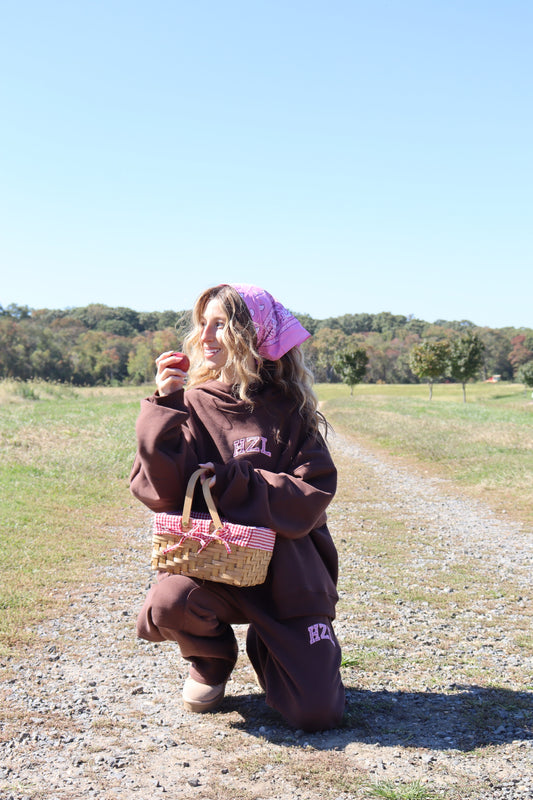 Woman wearing a brown mockneck hoodie and matching pants while kneeling outdoors with a woven basket