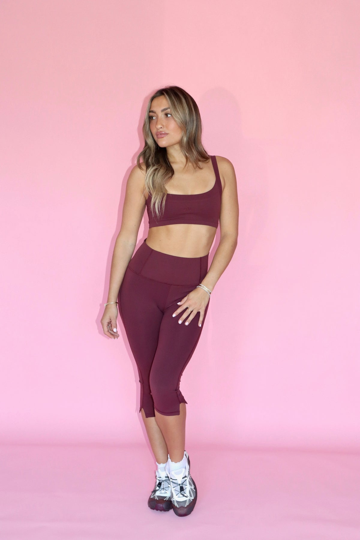Woman wearing a maroon crop top and matching capri leggings with sneakers against a pink background