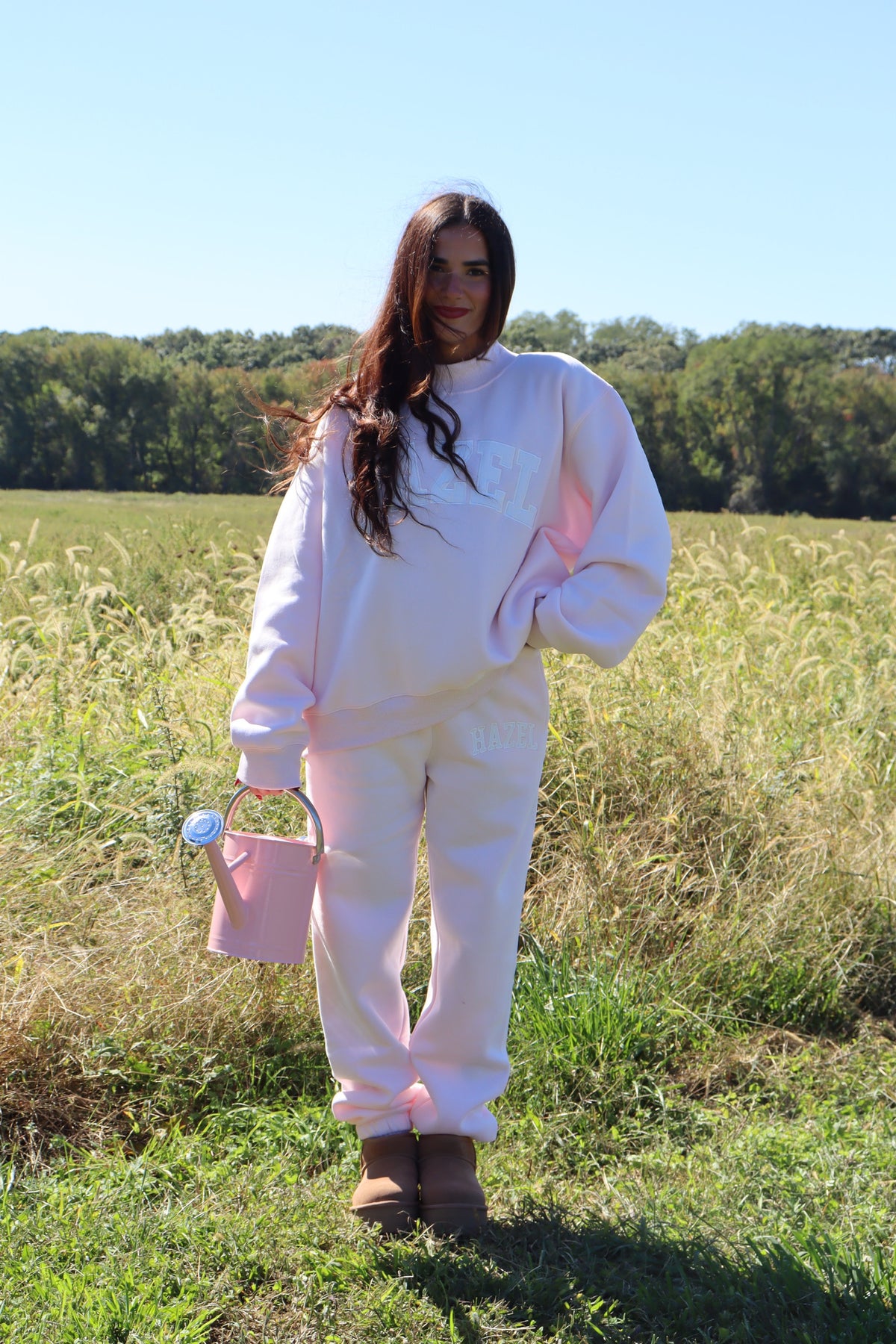 Woman wearing pale pink joggers and a matching sweatshirt in a sunny field with a pink handbag