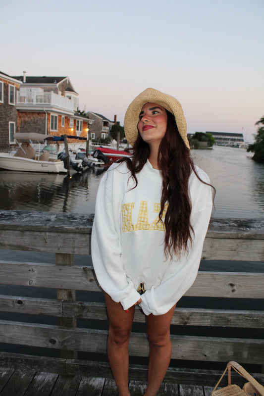 Young woman wearing a casual white sweatshirt with yellow lettering and a straw hat, standing by a waterfront dock at sunset.