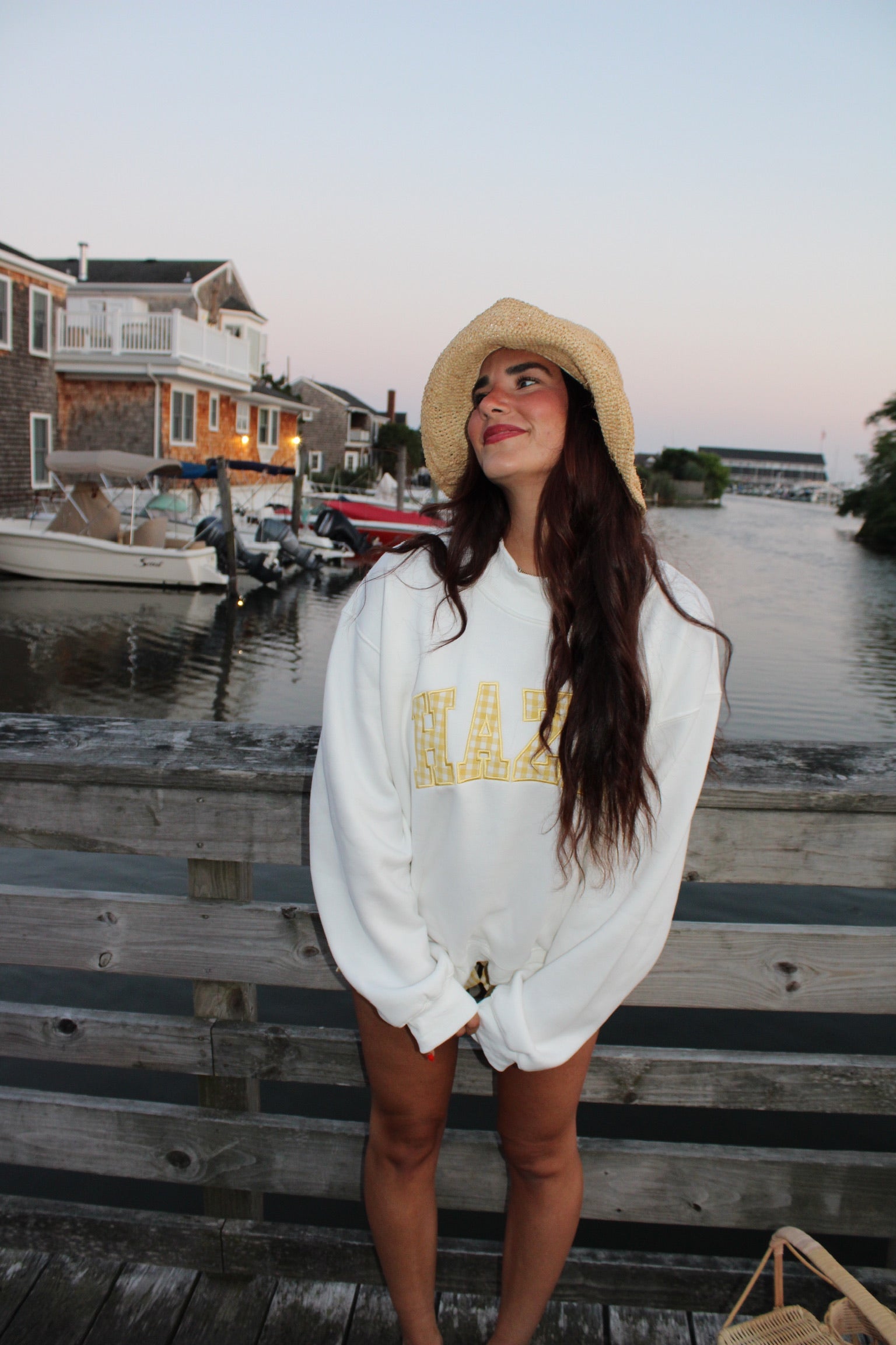 Young woman wearing a casual white sweatshirt with yellow lettering and a straw hat, standing by a waterfront dock at sunset.