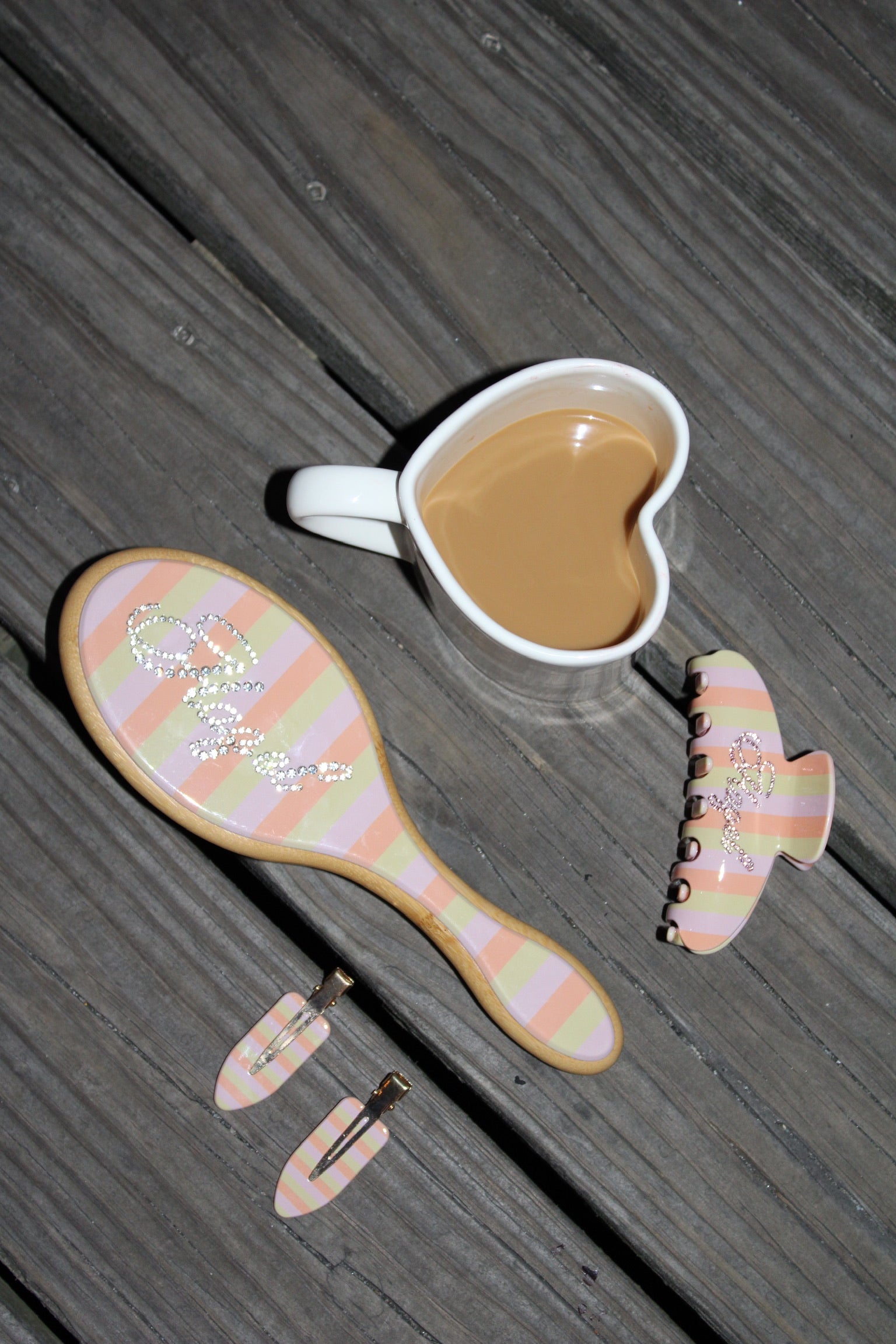 Heart-shaped cup of coffee next to pastel-striped hair accessories including a brush, claw clip, and hair pins with rhinestone details.