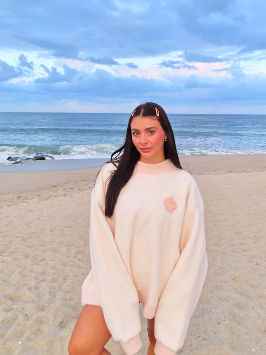 Woman wearing oversized light fleece pullover with small embroidered design on the beach at sunset