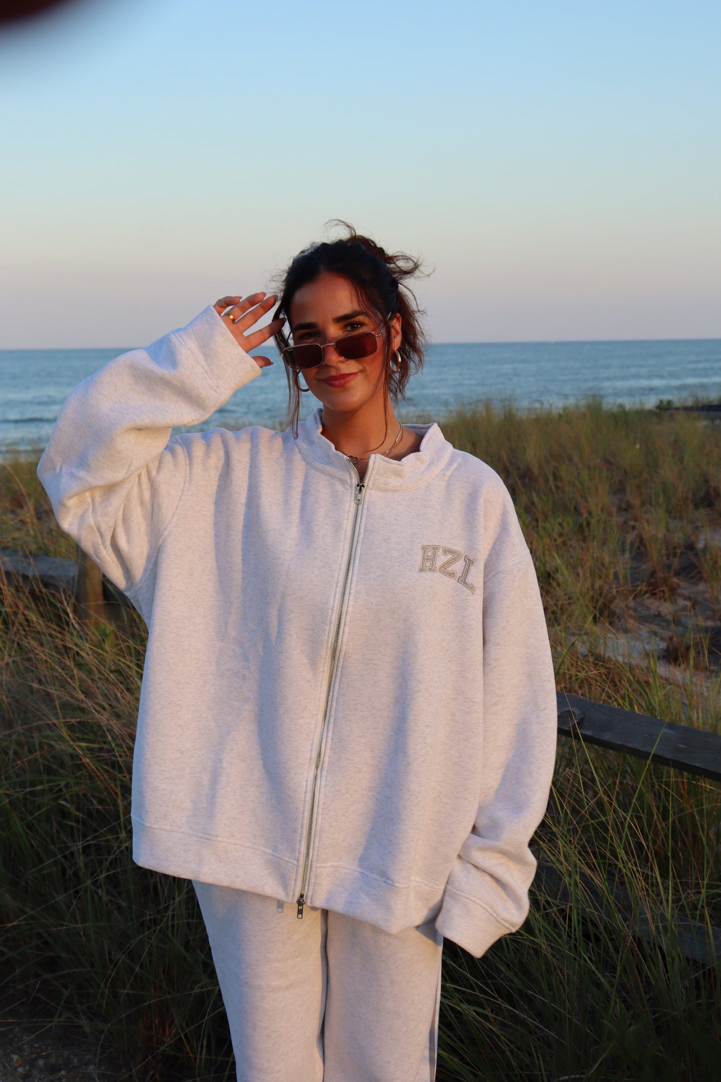 Woman wearing a cream HZL zip up sweatshirt and matching pants by beach dunes at sunset