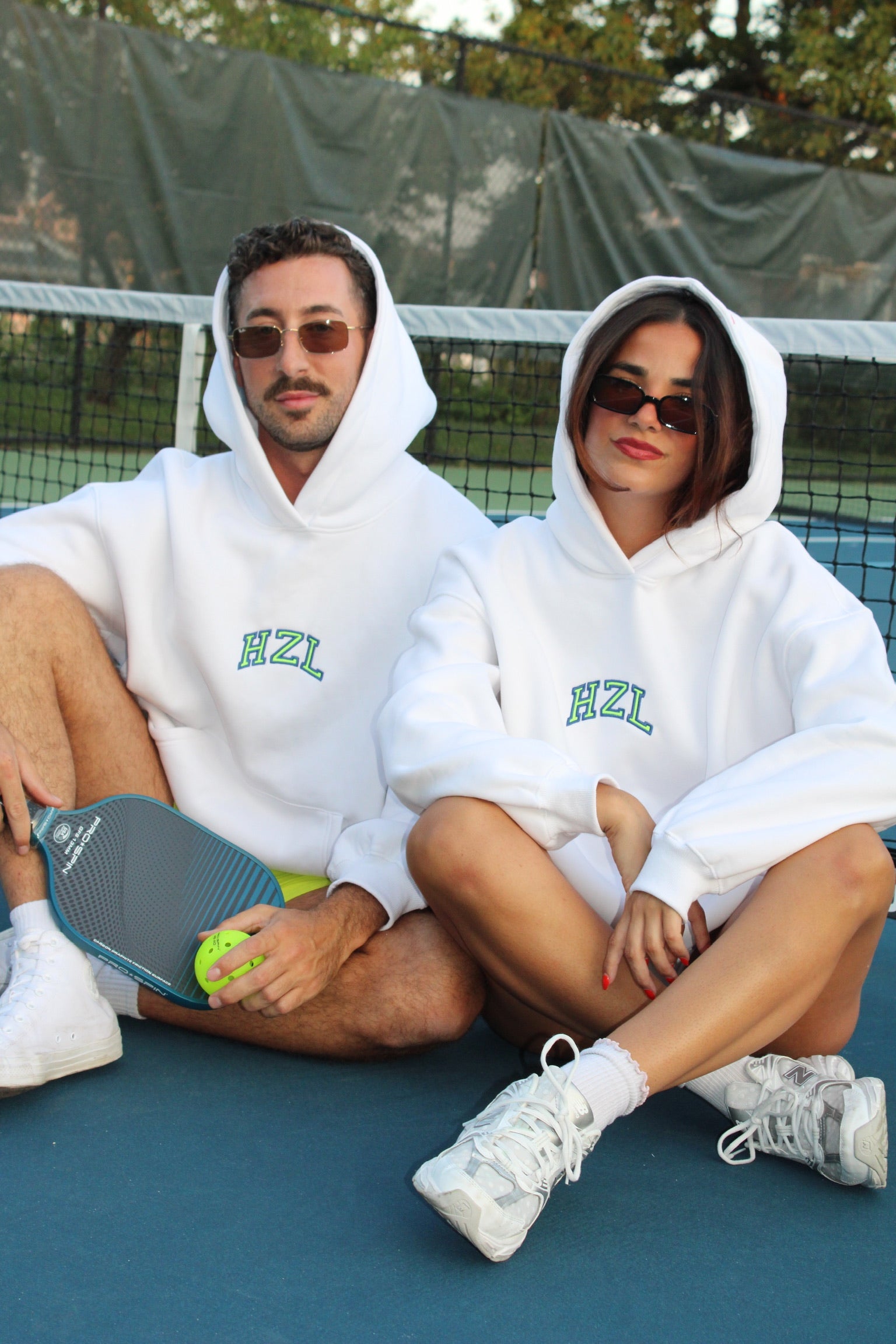 Two people wearing white HZL hoodies with hoods up sitting on a tennis court, each sporting sunglasses and casual sneakers.