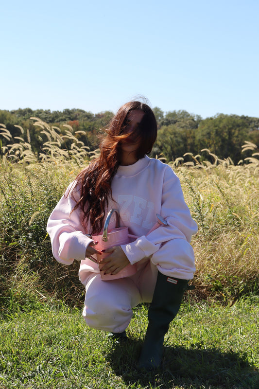 Woman wearing a pale pink mockneck sweatshirt and matching pants while holding a pink handbag in a grassy field