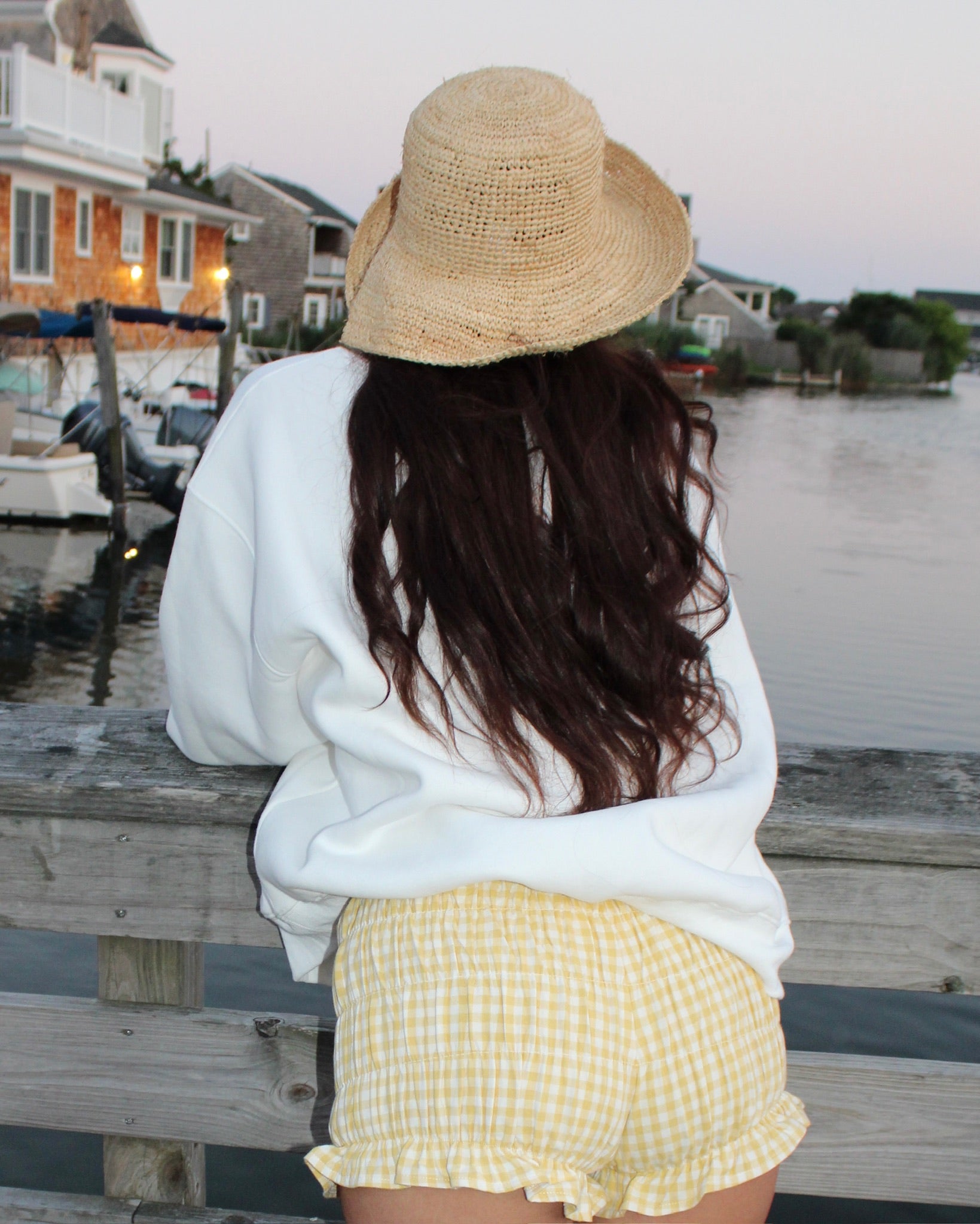 Woman wearing yellow gingham bloomers and a white sweatshirt, leaning on a wooden railing by the water at dusk.