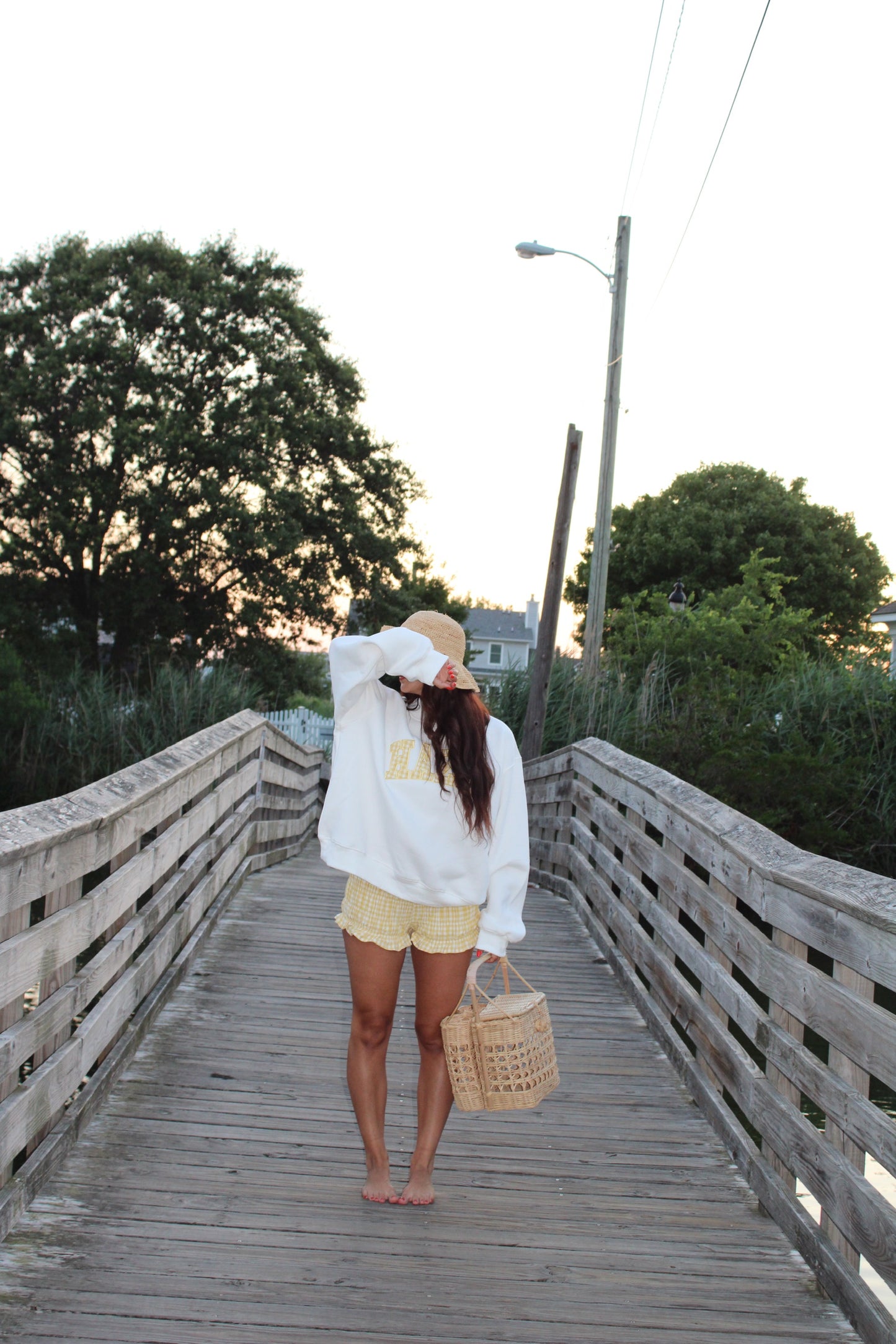 Woman standing barefoot on wooden boardwalk wearing a cozy white sweatshirt and yellow textured shorts, holding a woven basket bag.