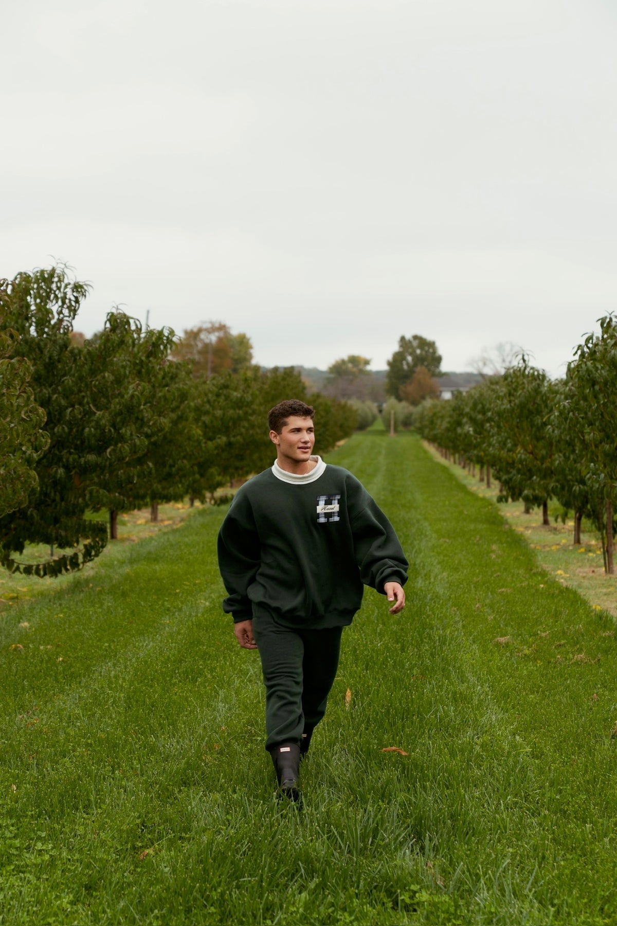 Model wearing green joggers and a matching sweatshirt walks through a grassy orchard