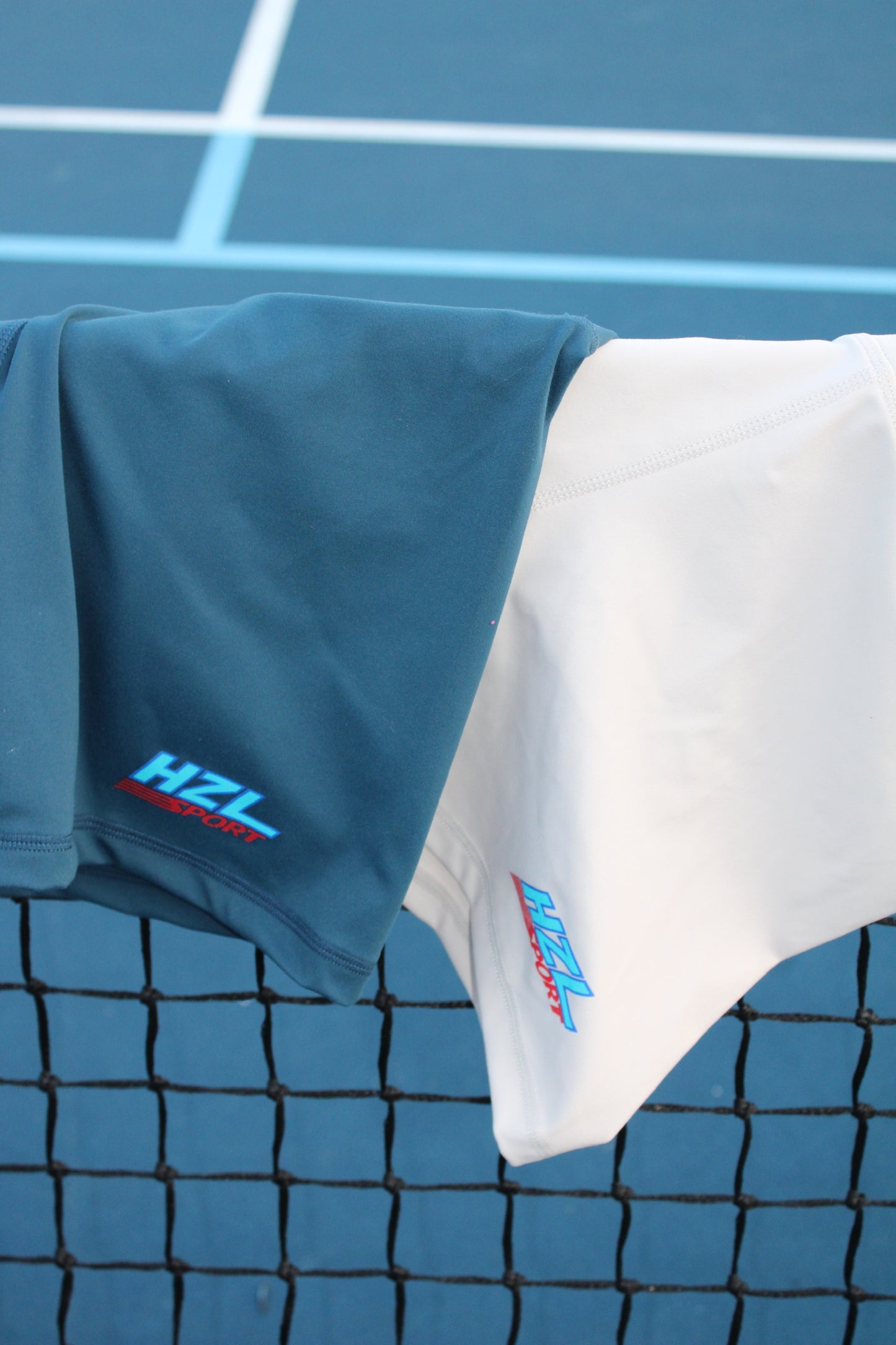 Two sporty shorts in dark blue and white hanging on a tennis net with a blue court background showing HZL Sport logos.
