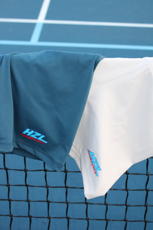 Two sporty shorts in dark blue and white hanging on a tennis net with a blue court background showing HZL Sport logos.