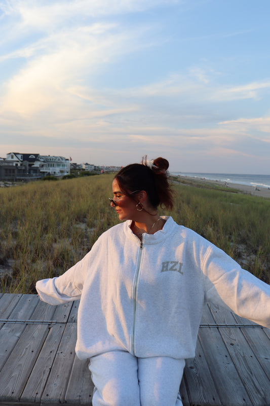 Woman wearing a light gray HZL zip-up sweatshirt and matching pants on a beach boardwalk