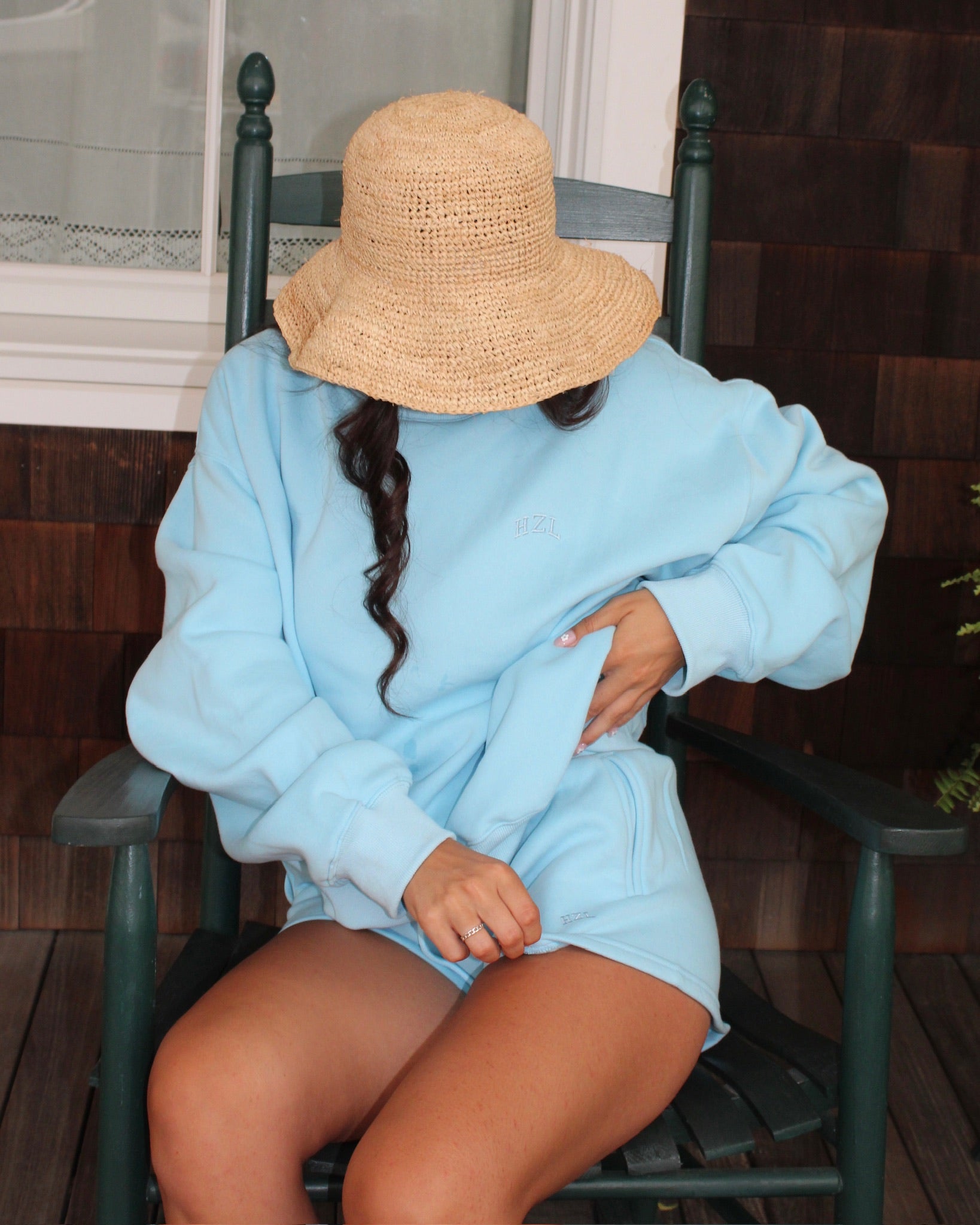 Person wearing a light blue cozy sweatshirt and matching shorts sitting on a dark wood rocking chair, with a wide-brimmed straw hat covering their face.