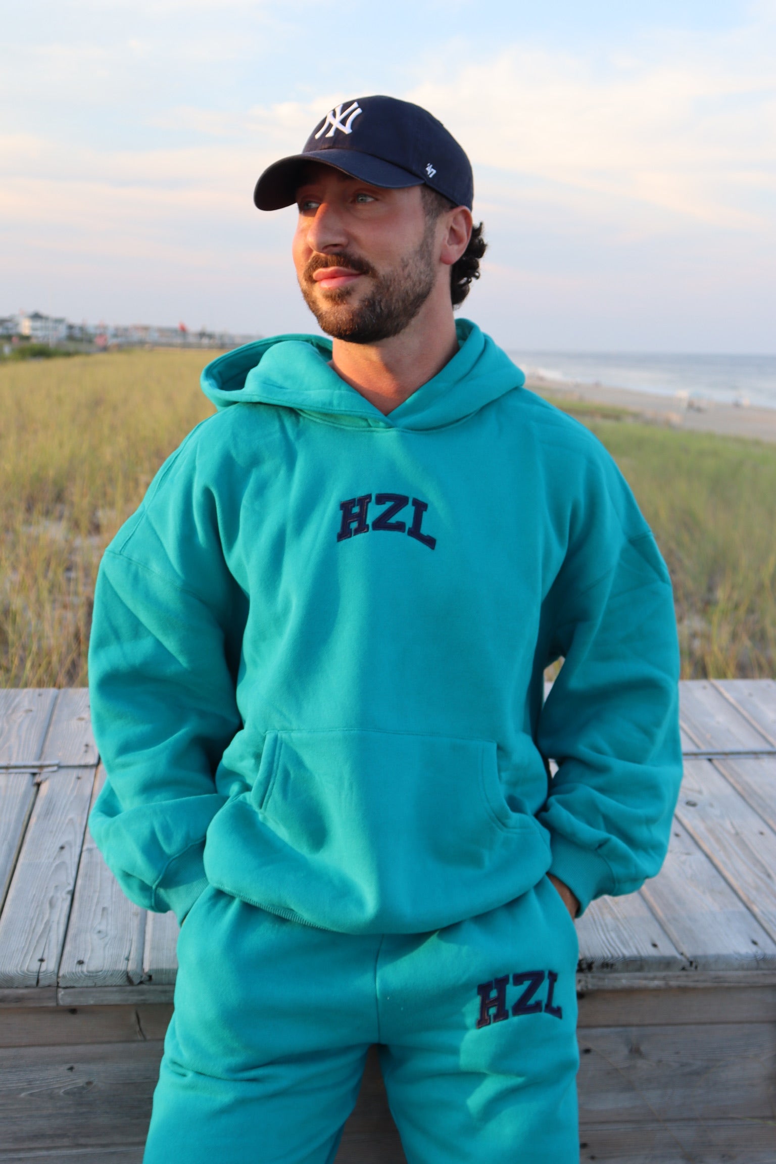 Man wearing a teal HZL hoodie and matching sweatpants on a beach boardwalk
