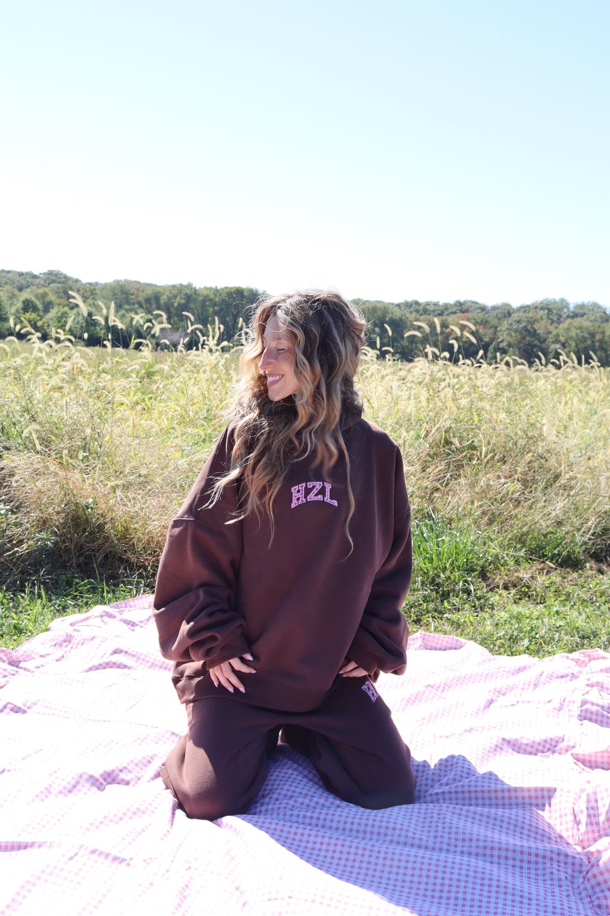 Woman kneeling on a picnic blanket in a field wearing a brown HZL mockneck hoodie