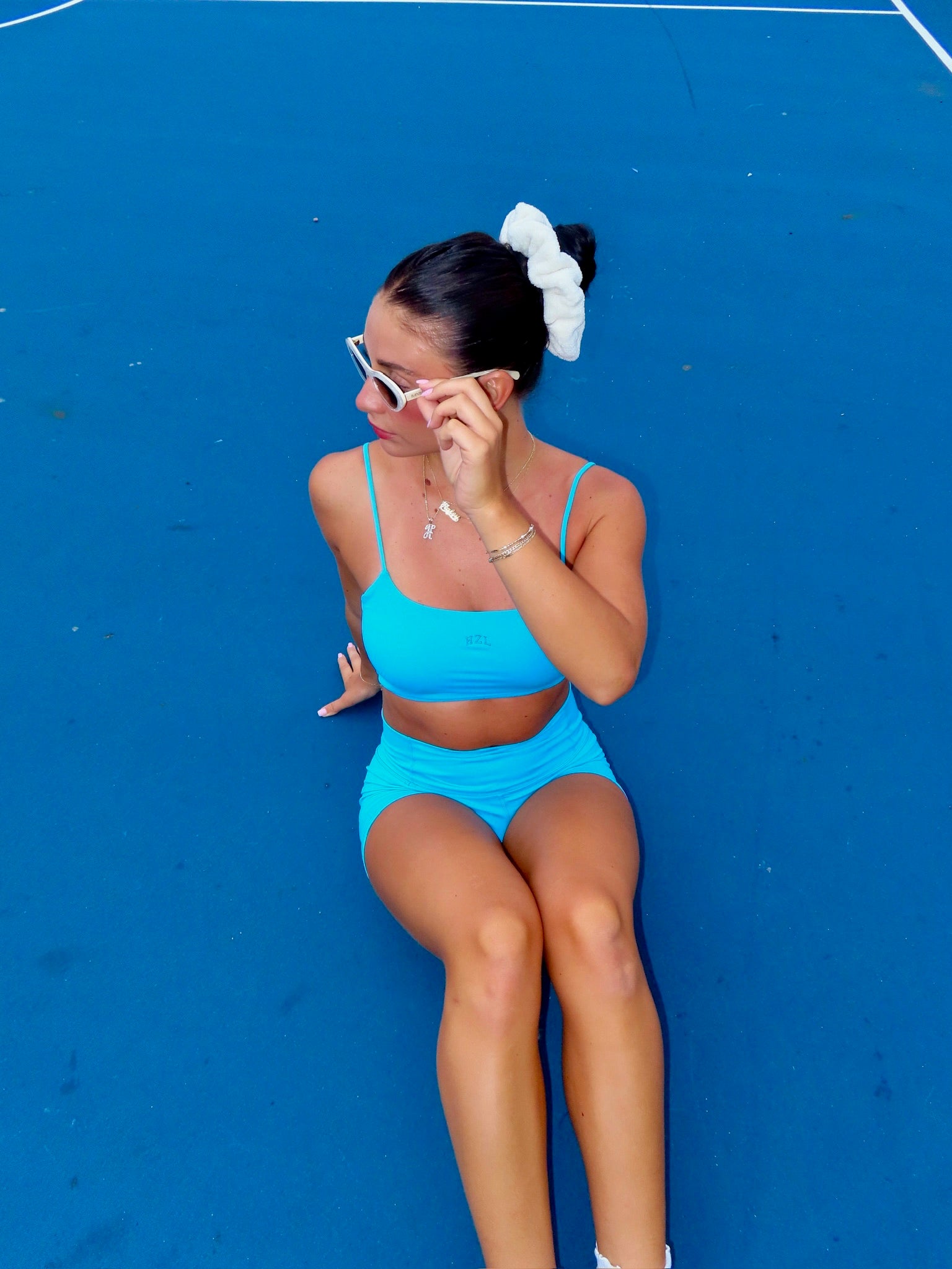 Woman sitting on a blue court wearing teal activewear shorts and a matching cropped top with sunglasses and a white scrunchie in her hair