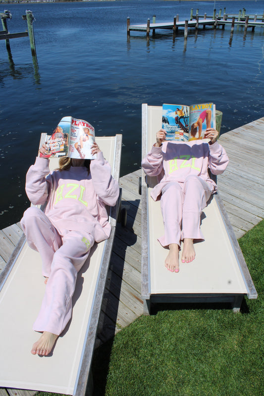 Two people relaxing on lounge chairs by the water, wearing pastel pink casual pants and sweatshirts while reading magazines.