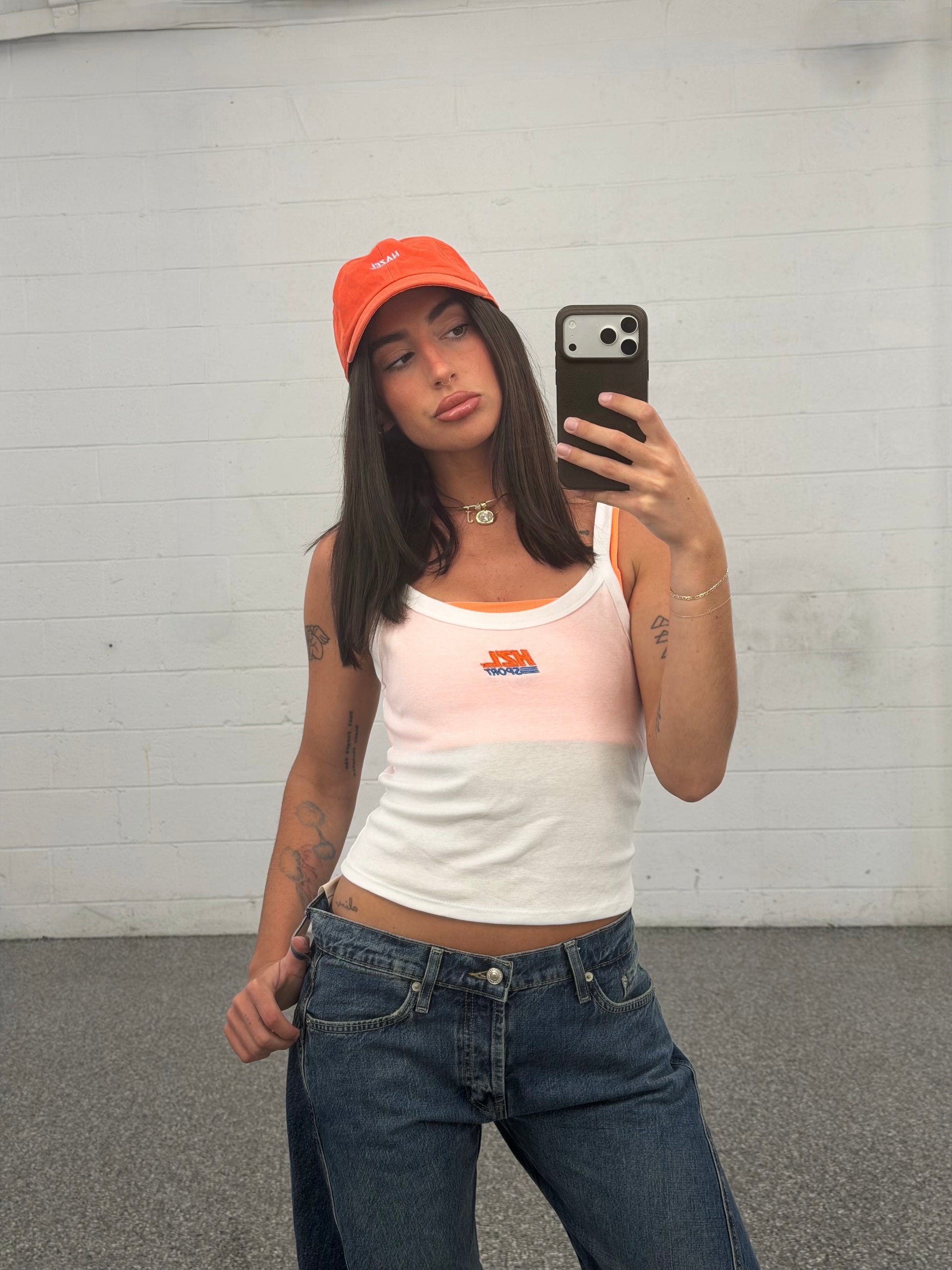 Woman wearing a white and light peach sporty tank top with a small logo, paired with blue jeans and an orange cap taking a mirror selfie.