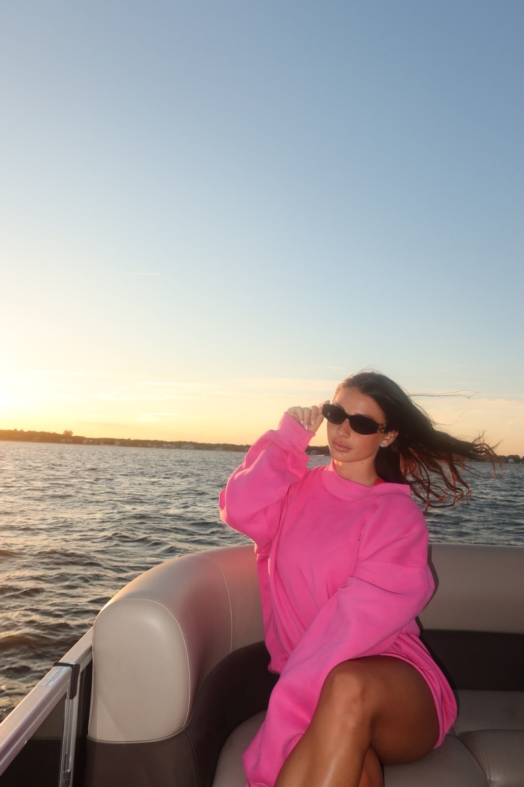 Woman wearing bright pink oversized mockneck dress sitting on a boat at sunset with sunglasses and windblown hair