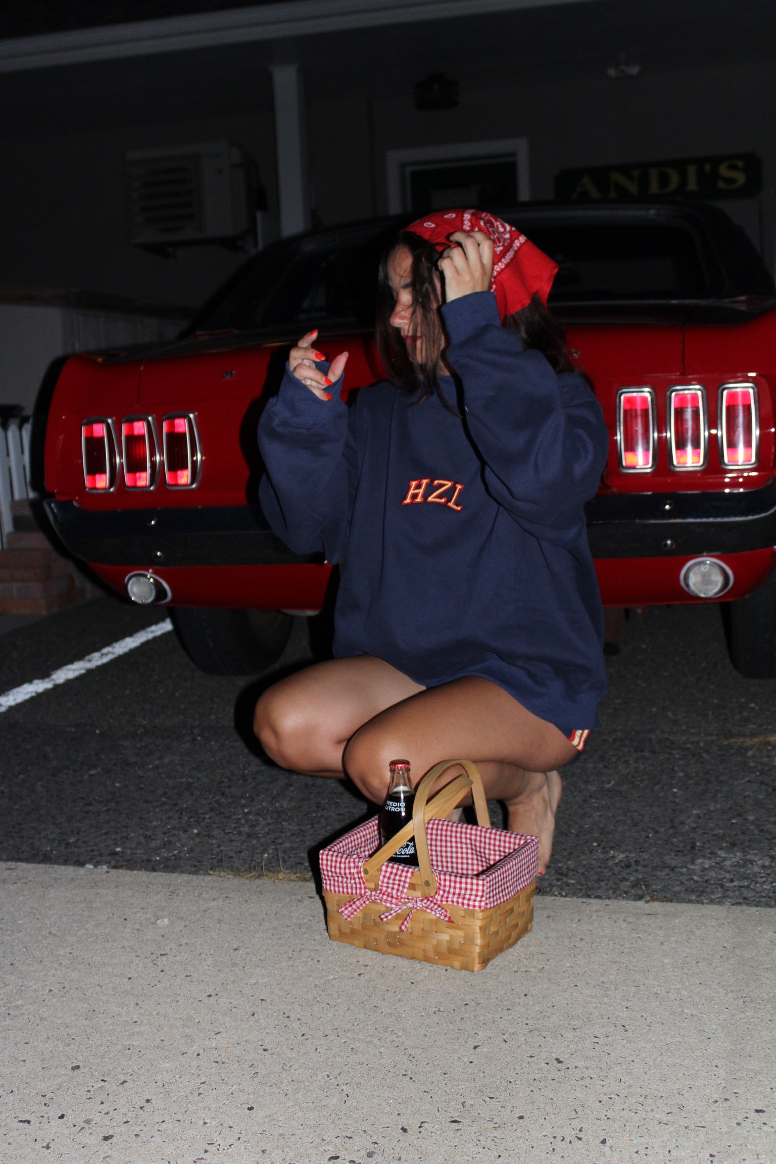 Young woman with red bandana squatting by a vintage red car, wearing a navy blue HZL sweatshirt and holding a picnic basket with a glass bottle inside.