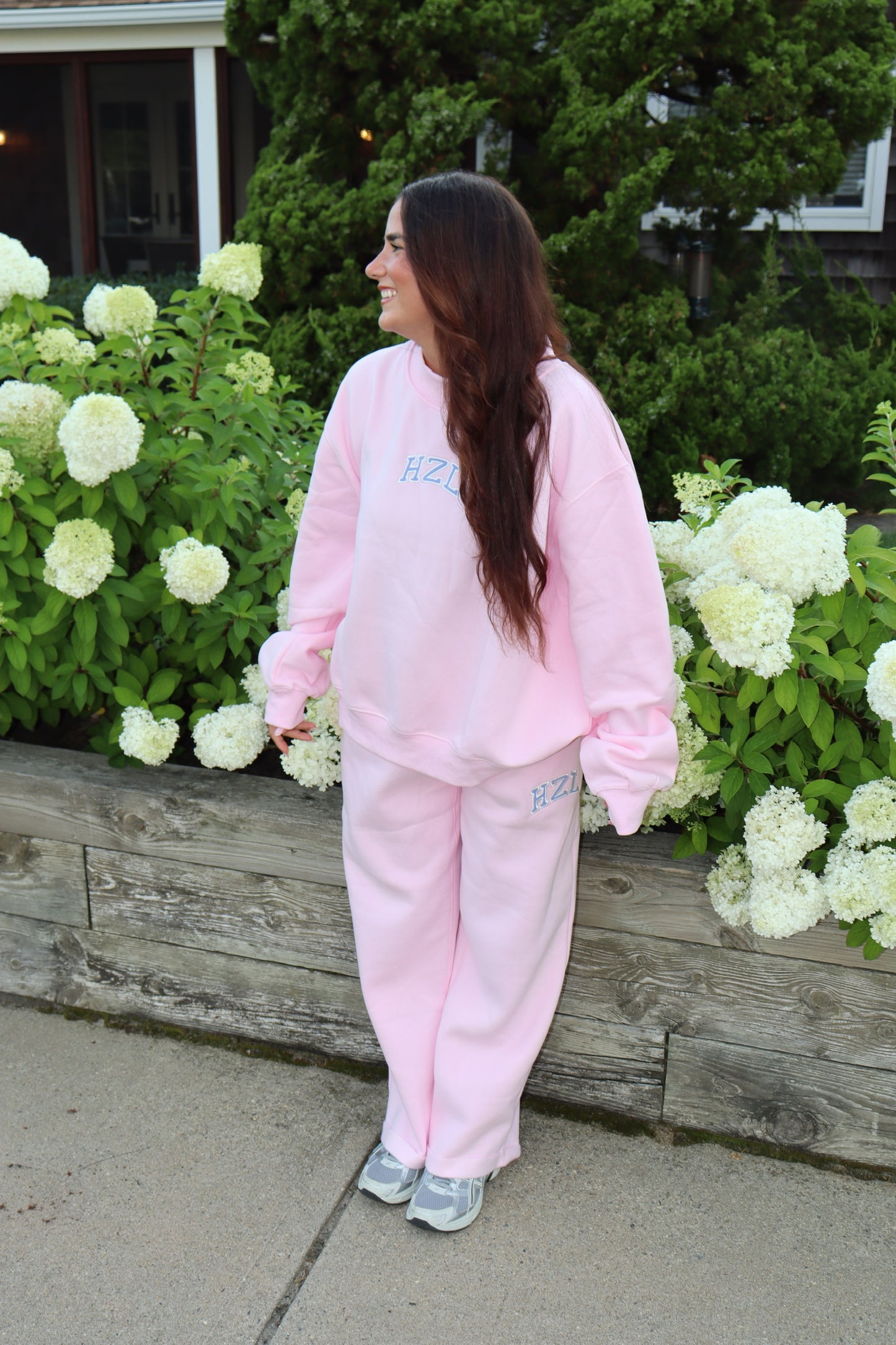 Woman wearing pink sweatpants and a matching sweatshirt beside blooming white hydrangeas