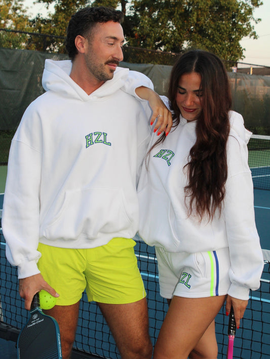Two people wearing white hoodies with green "HZL" logo standing on a tennis court, one wearing bright yellow shorts and the other white sporty shorts with blue and green stripes.