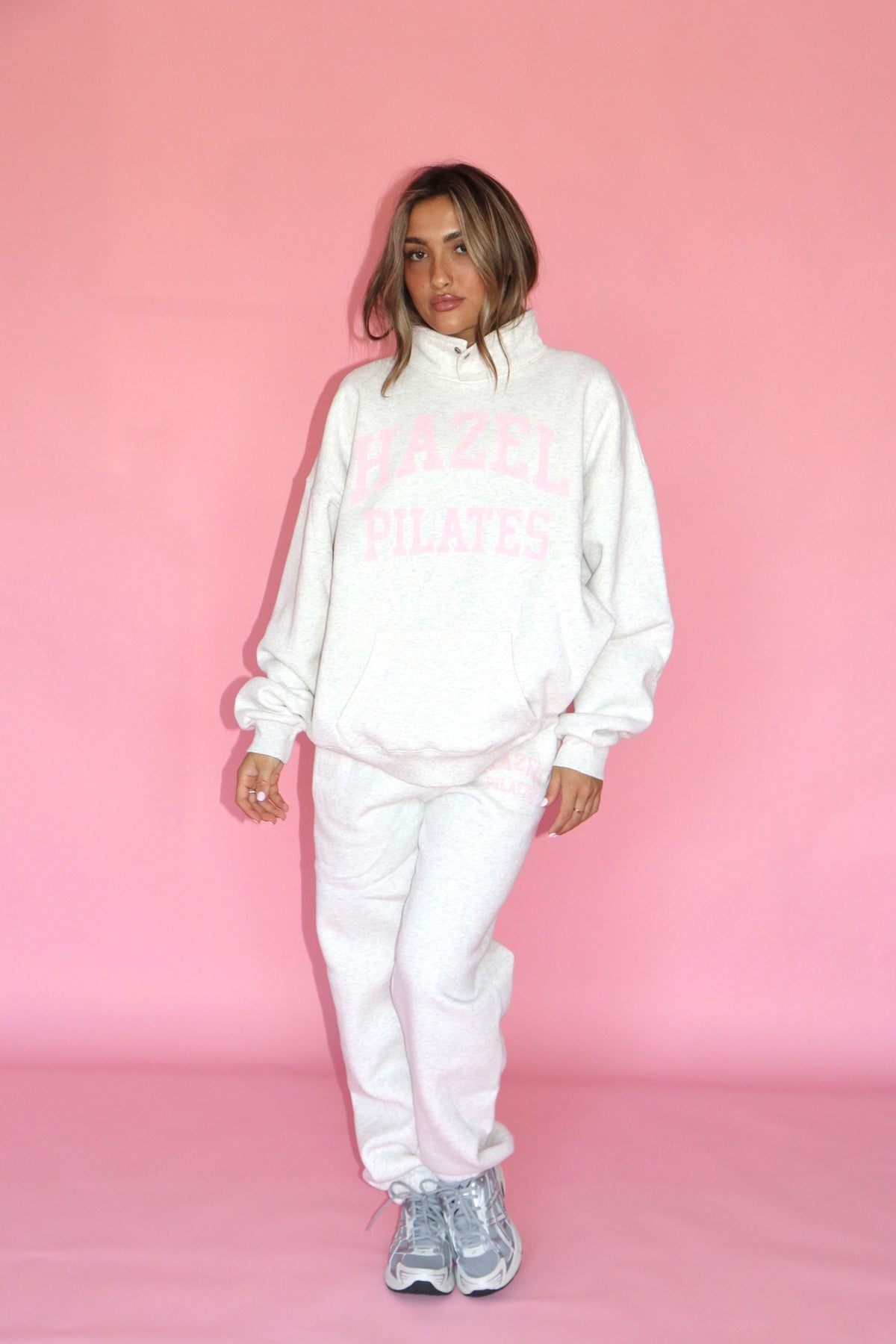 Model wearing a white Pilates sweatshirt and matching sweatpants with sneakers against a pink backdrop