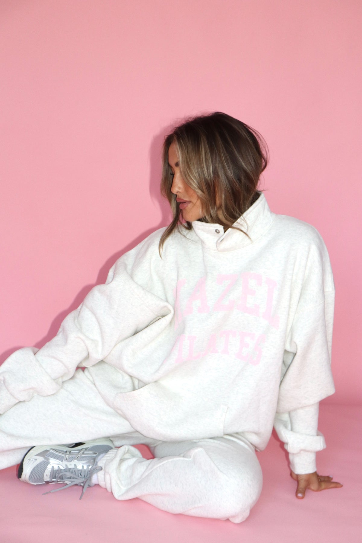 Woman seated in a white sweatshirt and matching sweatpants against a pink backdrop