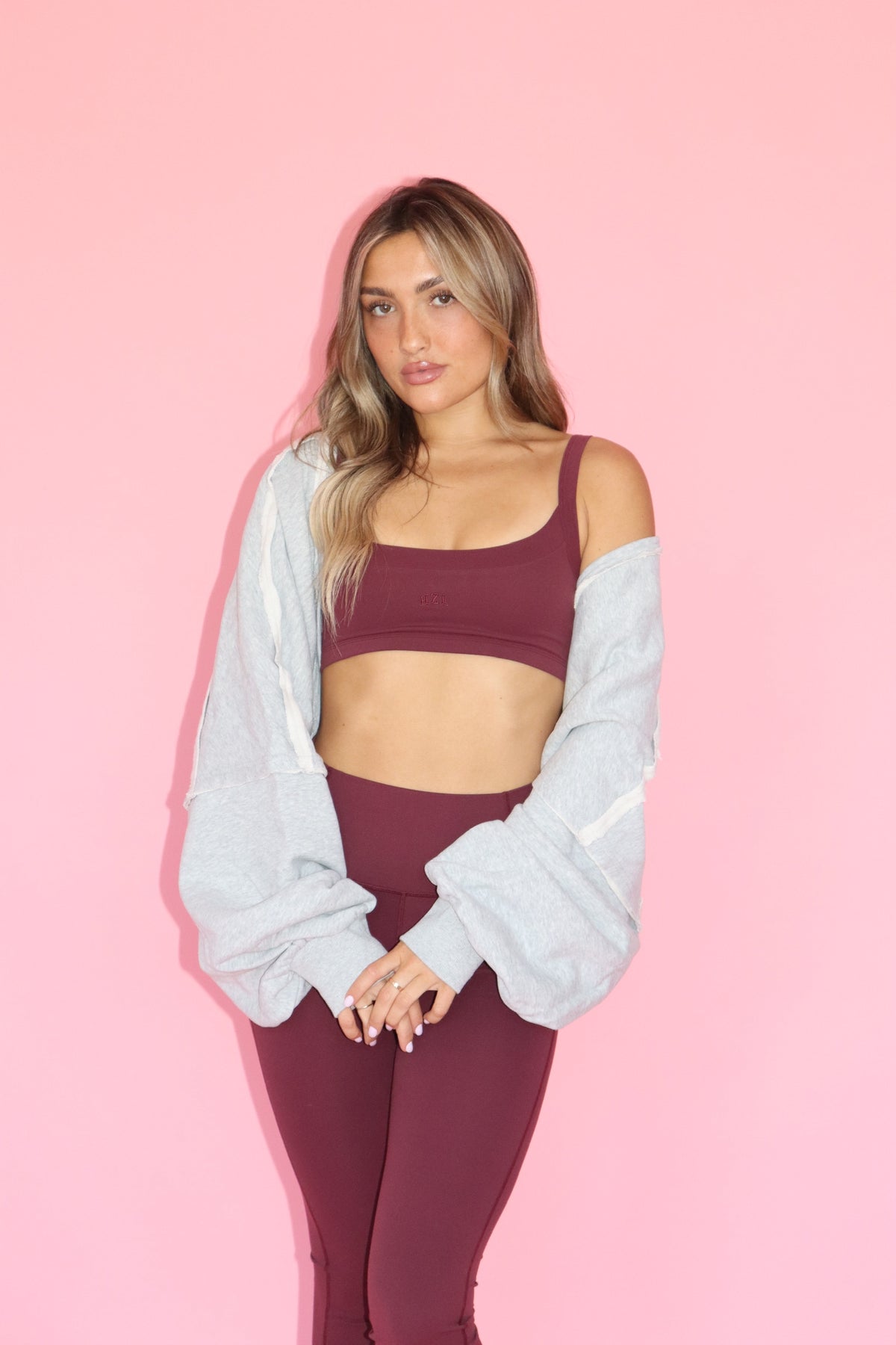 Model wearing a grey warm up shrug over burgundy activewear against a pink background