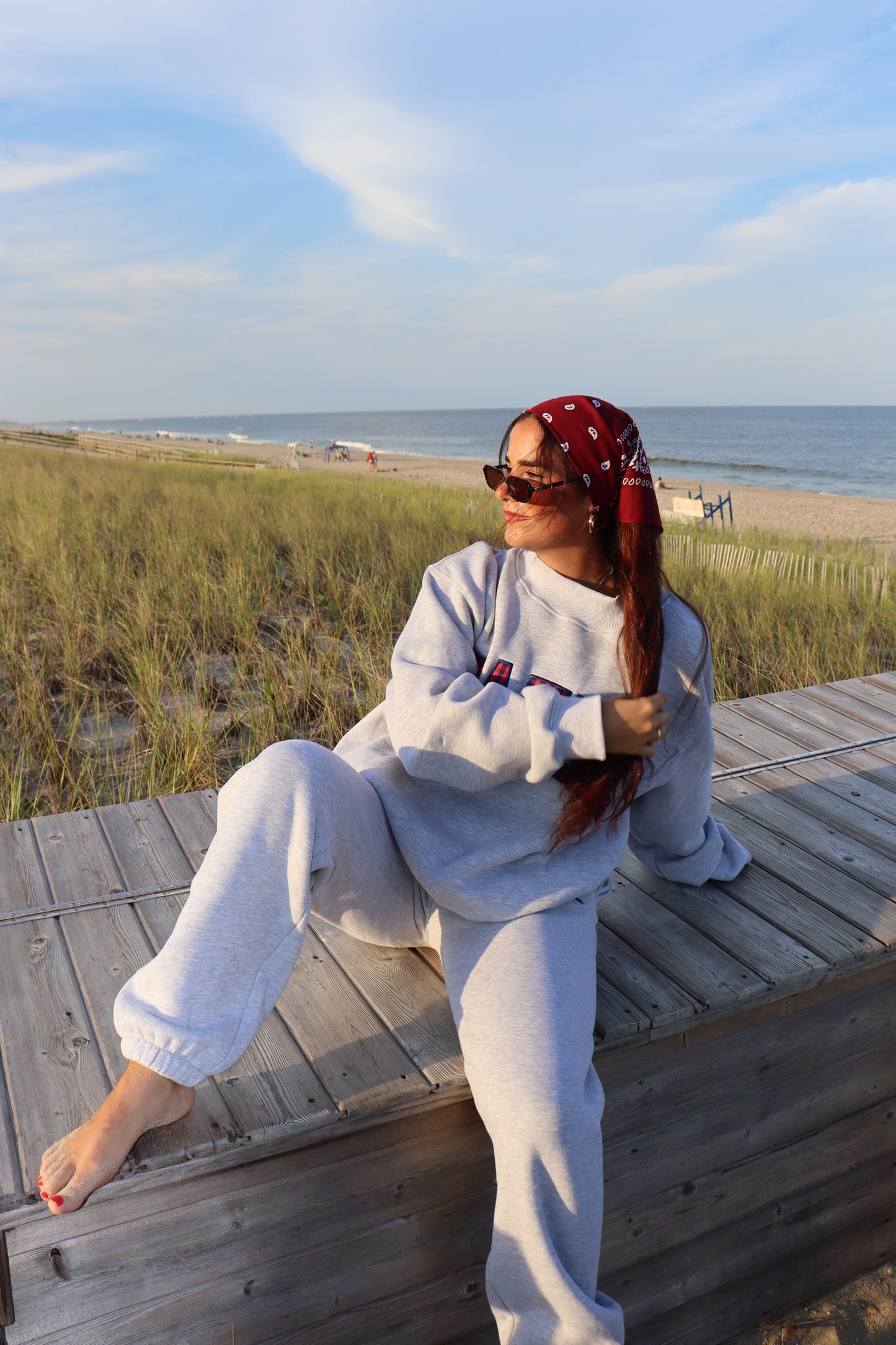 Woman wearing gray joggers and a matching sweatshirt sits on a boardwalk beside the beach