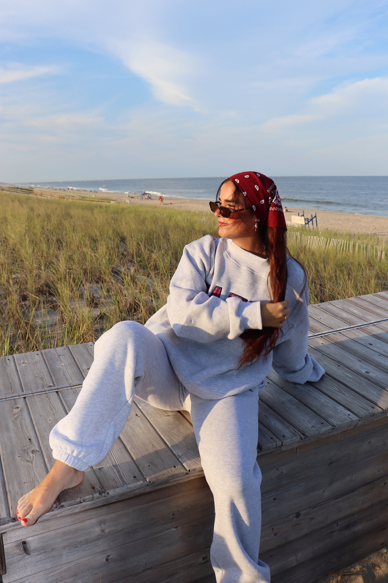 Woman wearing gray joggers and a matching sweatshirt sits on a boardwalk beside the beach