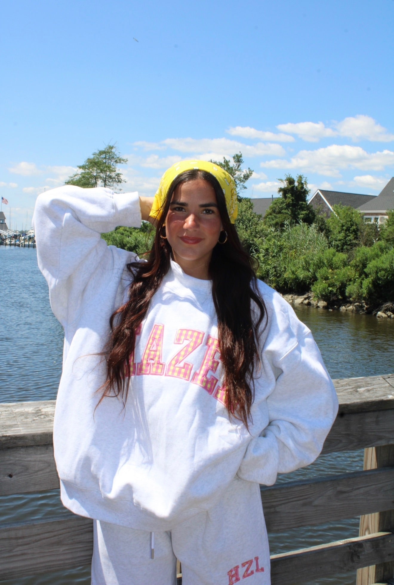 Young woman wearing a casual white sweatshirt and matching pants with bright lettering, posing outdoors by a marina on a sunny day