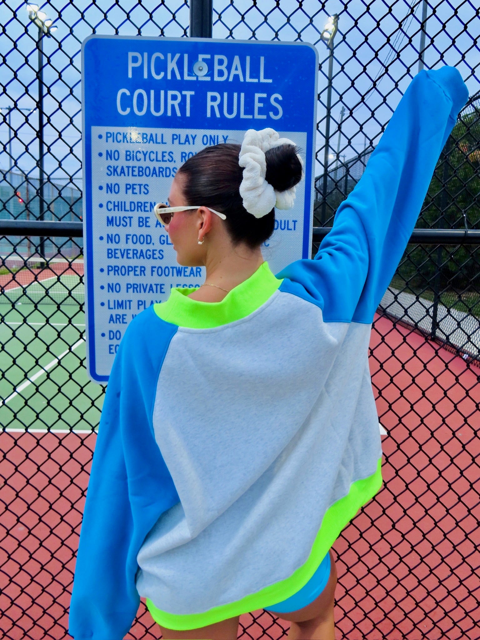 Woman with hair in a scrunchie wearing a color-block sweatshirt with blue sleeves and neon green details near a pickleball court sign