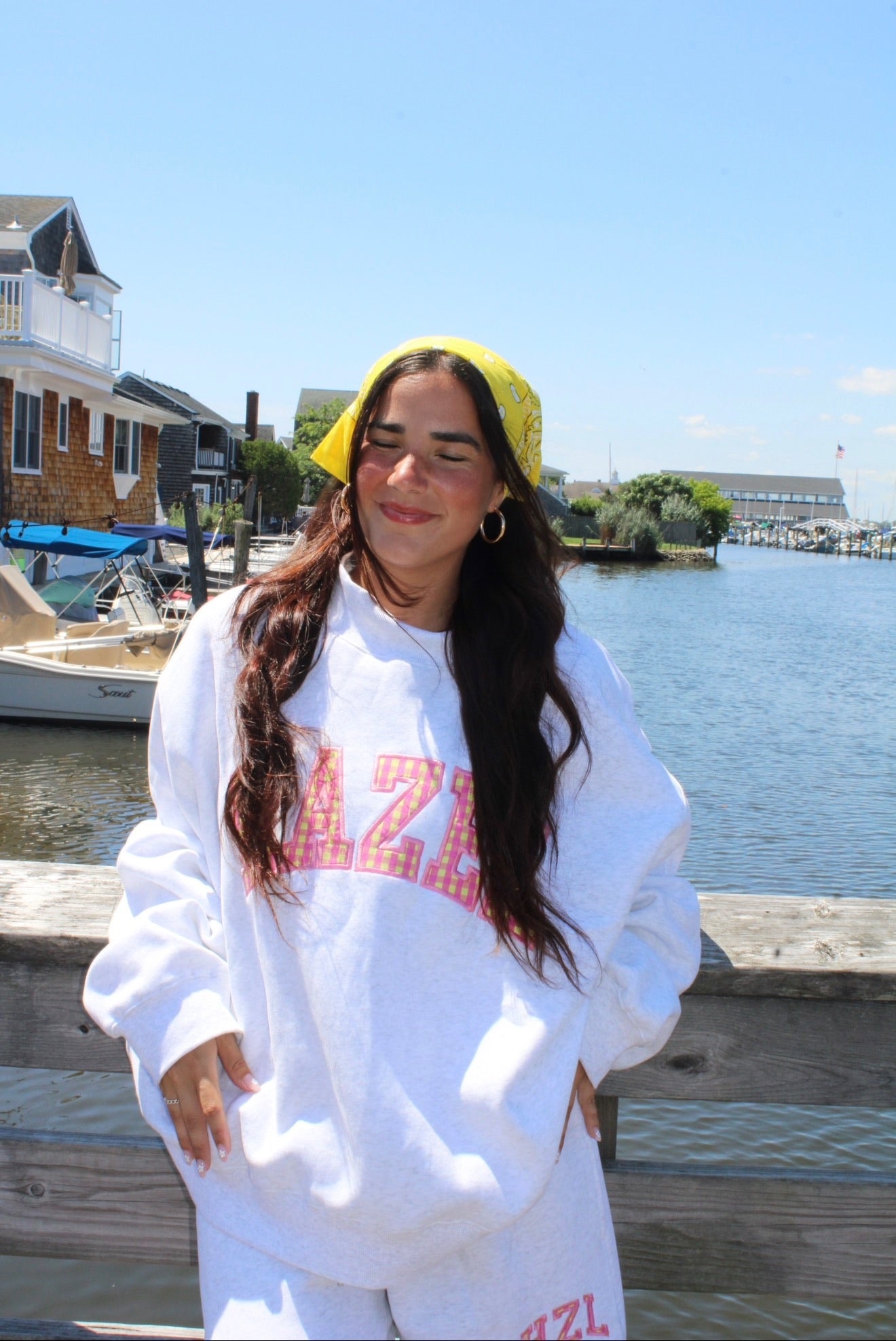 Young woman with long dark hair wearing a white oversized sweatshirt and matching shorts, smiling and standing by a wooden railing near a waterfront with boats and houses in the background.
