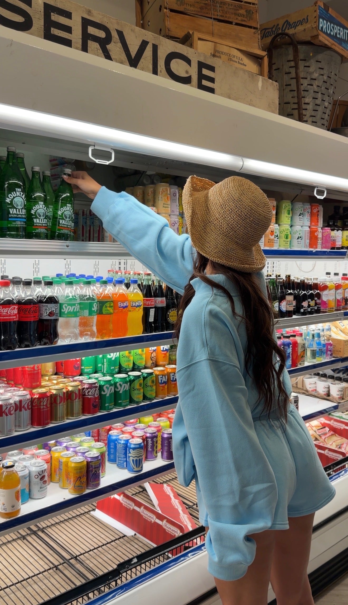 Woman wearing light blue shorts and oversized sweatshirt reaching for a drink in a grocery store while wearing a woven bucket hat