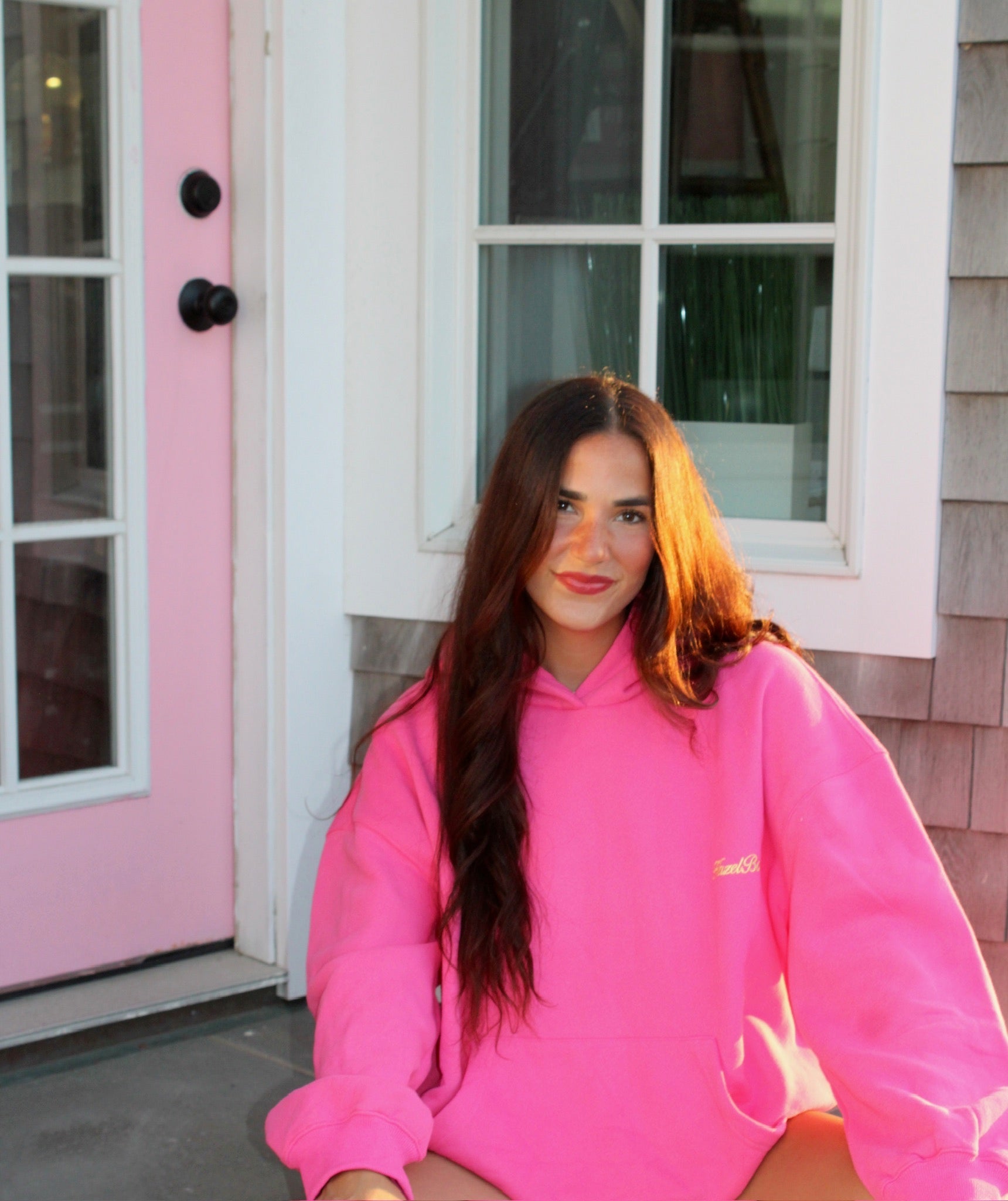 Young woman wearing a bright pink casual hoodie sitting outside a house with a pink door in soft sunlight.