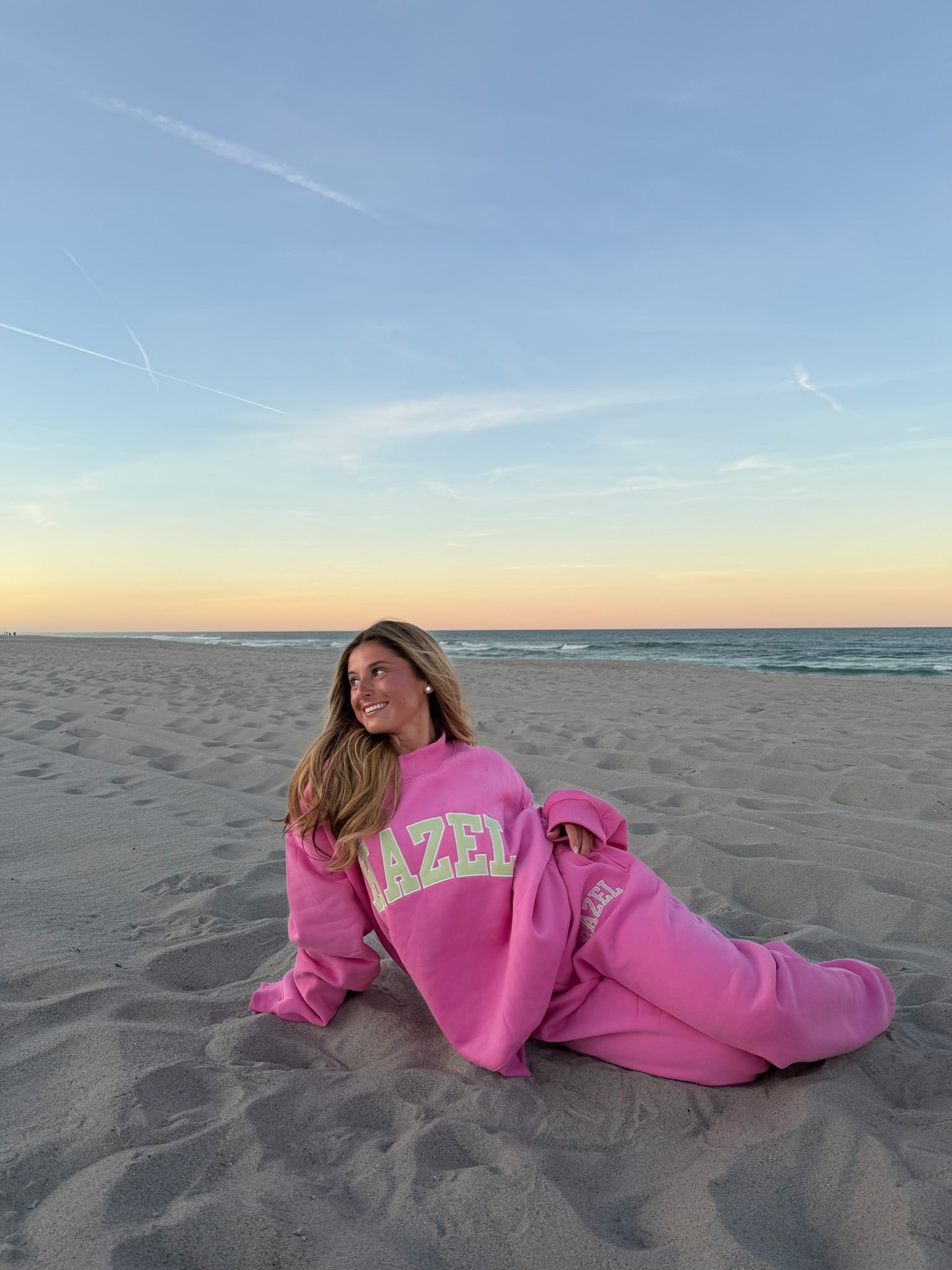 Woman relaxing on the beach at sunset wearing a cozy pink Hazel Boutique sweatshirt and matching sweatpants