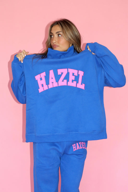 Woman wearing a bright blue high neck sweatshirt with pink HAZEL lettering and matching pants