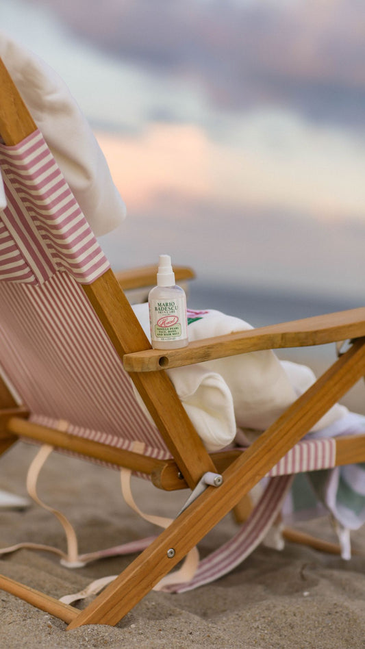 A wooden beach chair with a pink and white striped fabric seat holding a spray bottle of Vanilla Pearl Mist against a sandy shore backdrop.