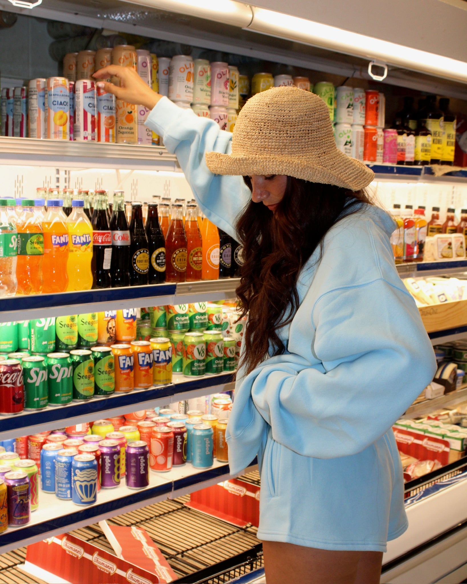 Woman wearing light blue oversized sweatshirt and shorts reaching for a beverage in a grocery store cooler while wearing a tan woven hat