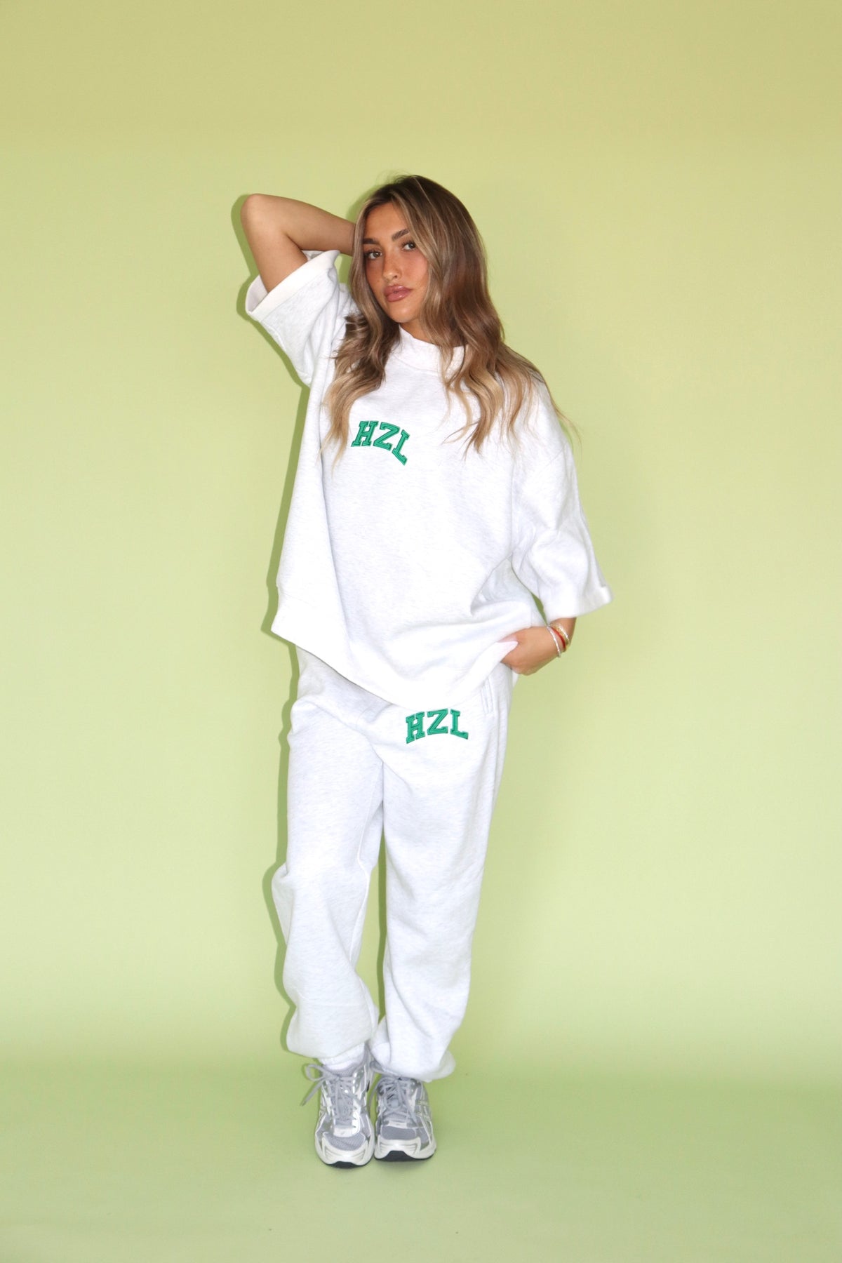 Woman wearing a white HZL short sleeve mockneck top with matching jogger pants and sneakers against a green backdrop