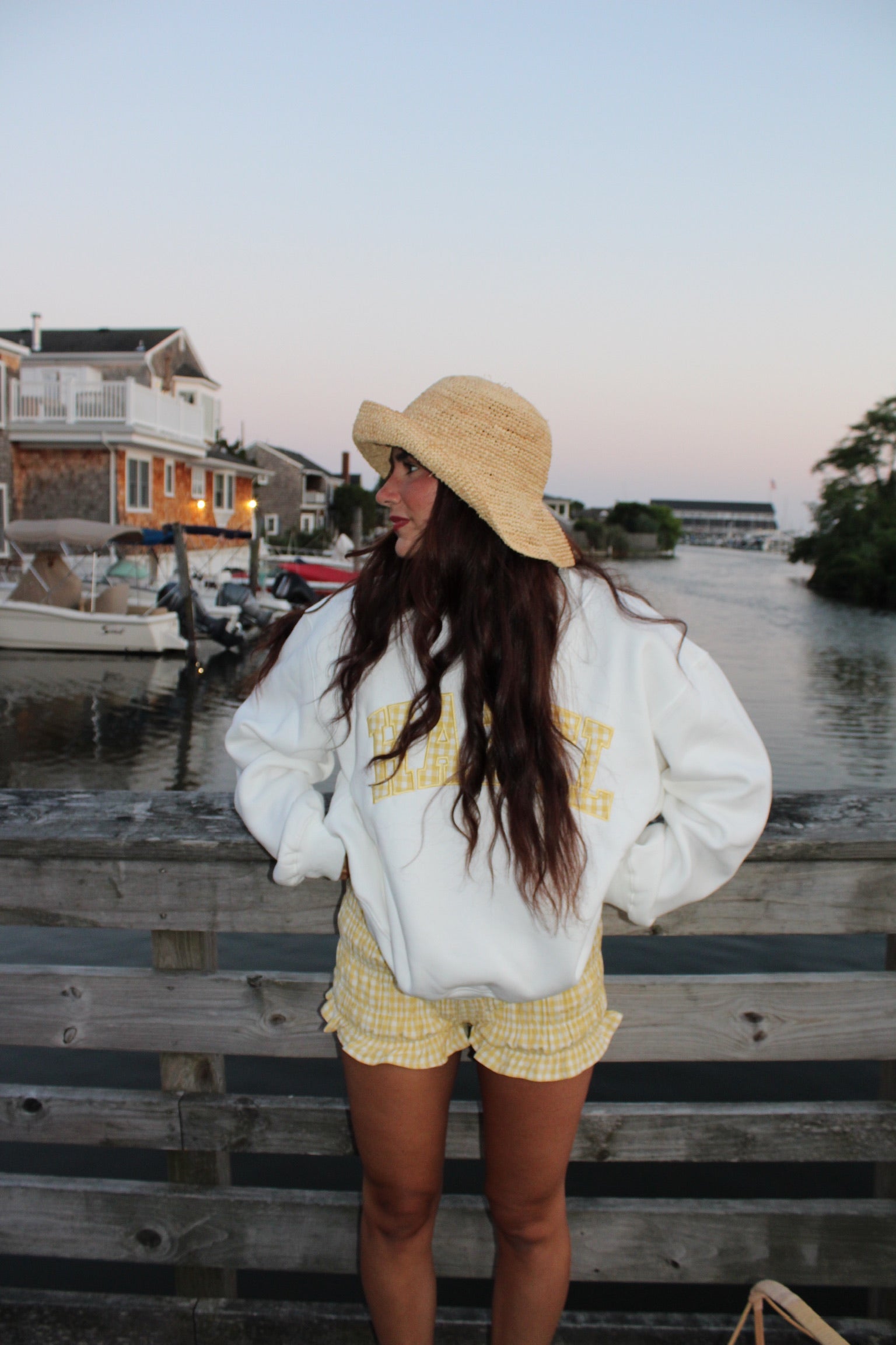 Woman wearing yellow gingham ruffled shorts and a straw hat stands by a wooden pier near water at dusk
