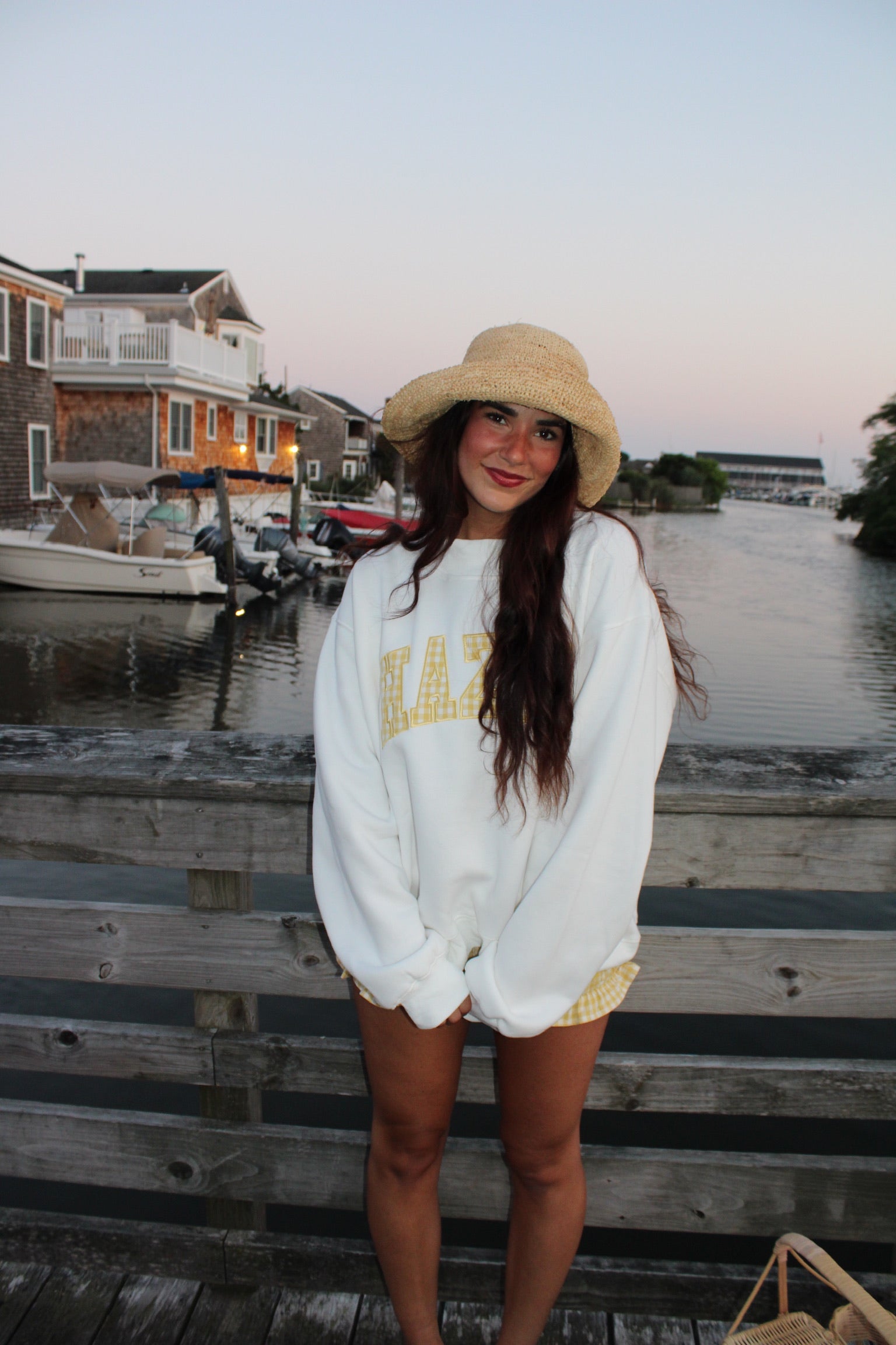 Young woman wearing a white oversized sweatshirt and straw hat standing on a wooden dock by the water at sunset