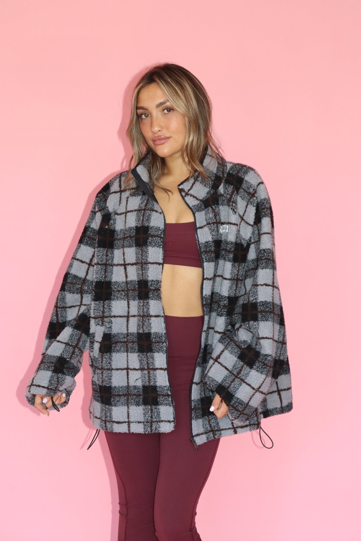 Woman wearing a gray and black plaid fleece jacket over a burgundy top and leggings against a pink backdrop