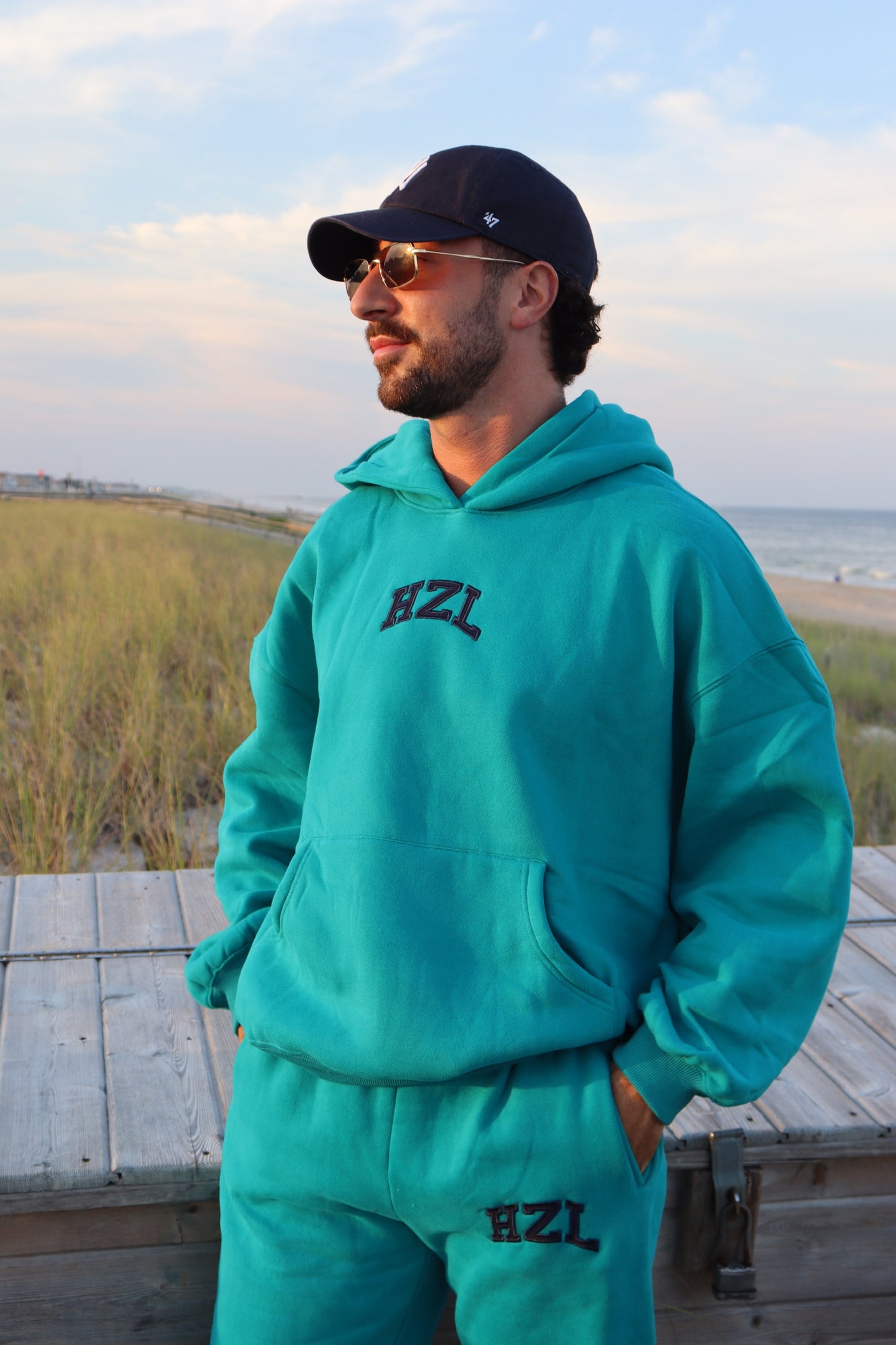 Man wearing a turquoise HZL hoodie and matching sweatpants on a beach boardwalk