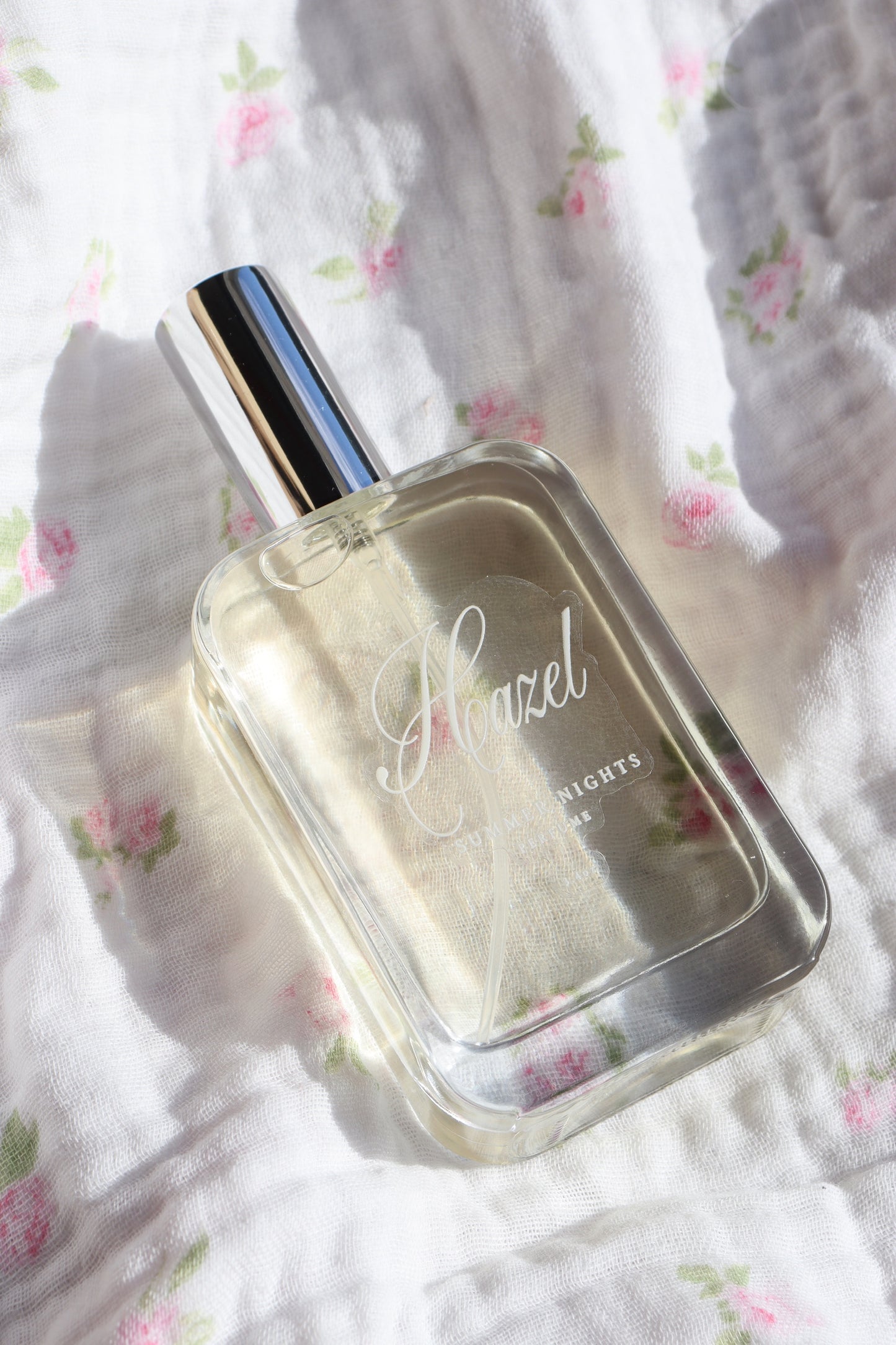 Hazel Summer Nights perfume bottle resting on floral white fabric in soft sunlight