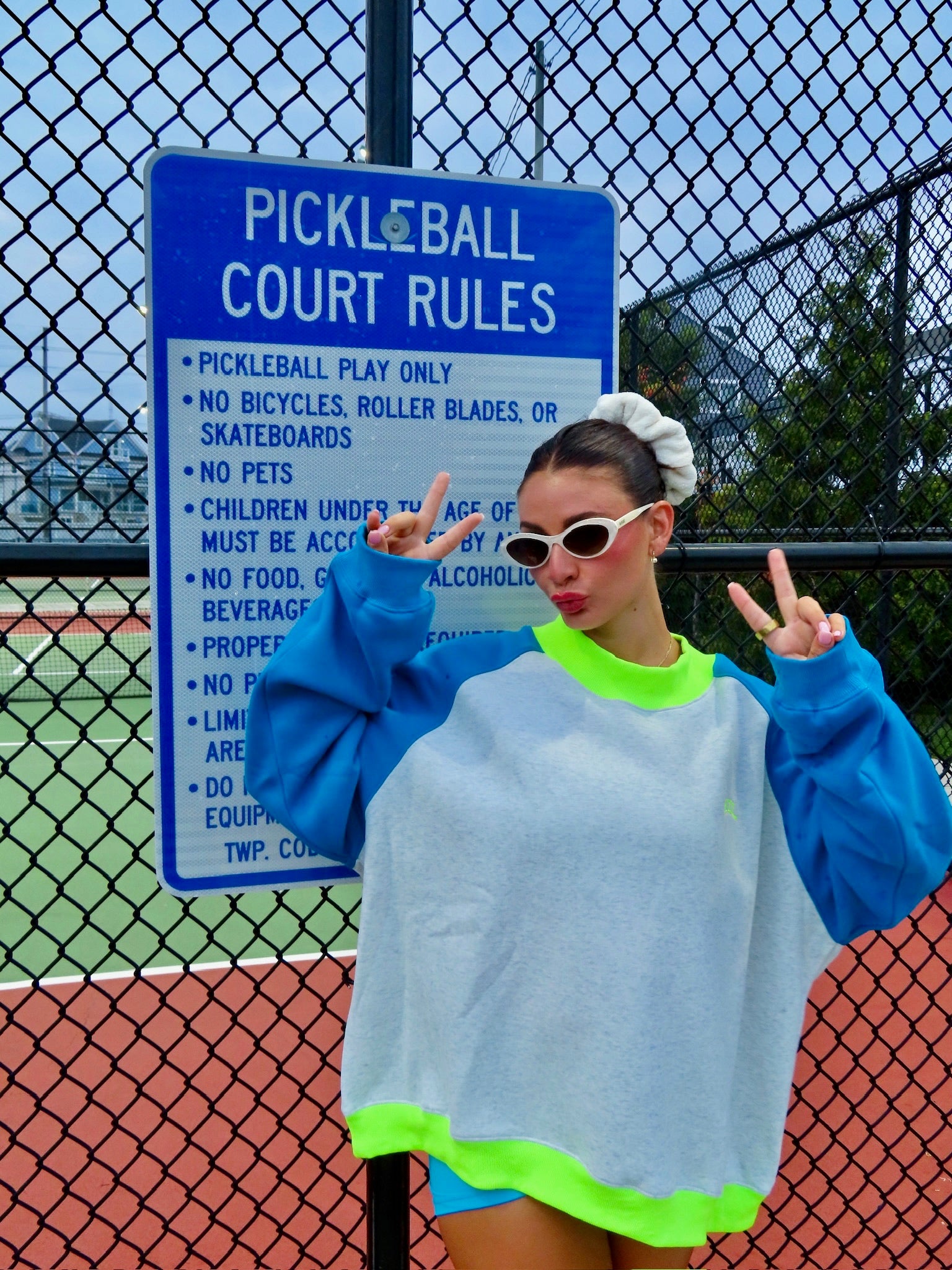 Woman wearing a colorful mockneck sweatshirt and white sunglasses posing with peace signs in front of a pickleball court fence