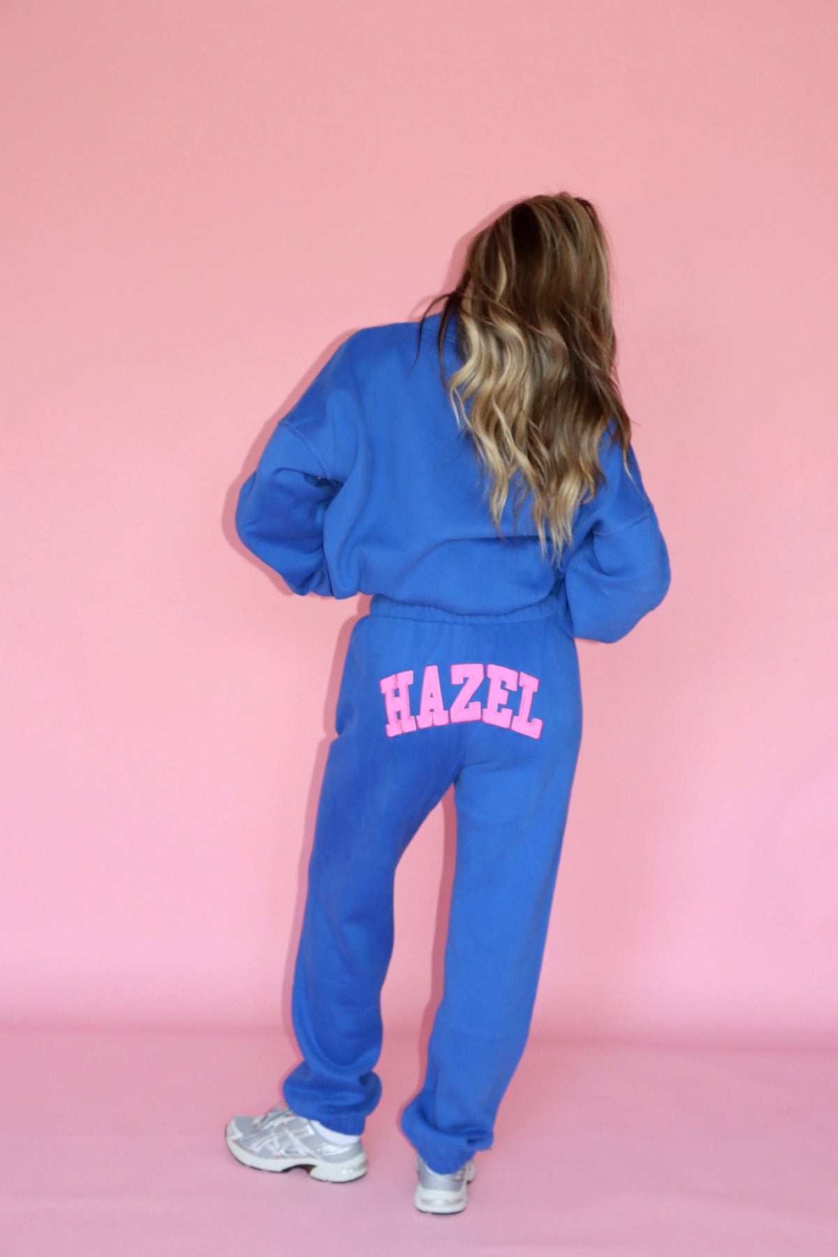 Back view of a model wearing blue joggers with pink HAZEL lettering and a matching sweatshirt against a pink backdrop
